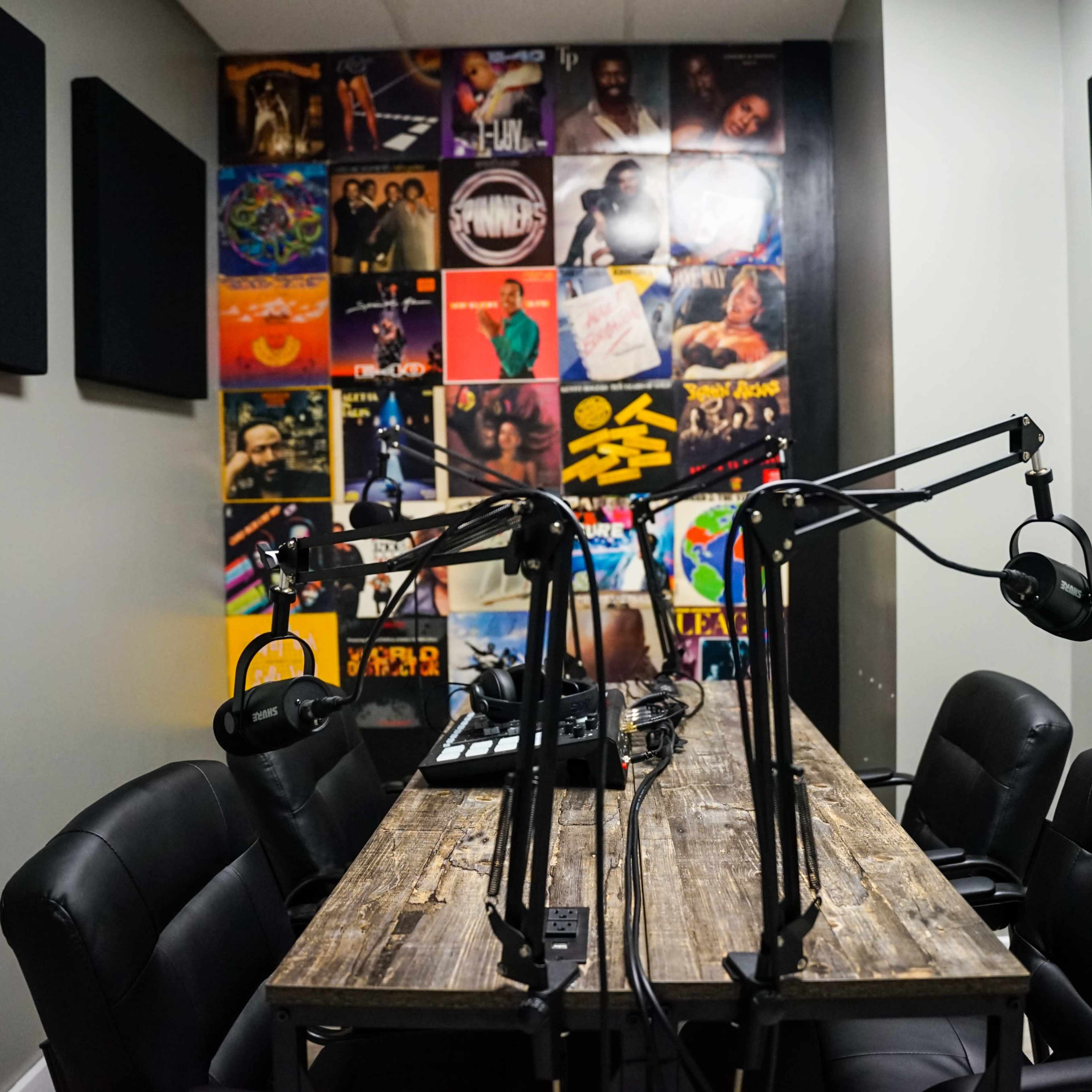 A small recording studio features a wooden table surrounded by black chairs and two microphone setups, with a wall covered in colorful album art.