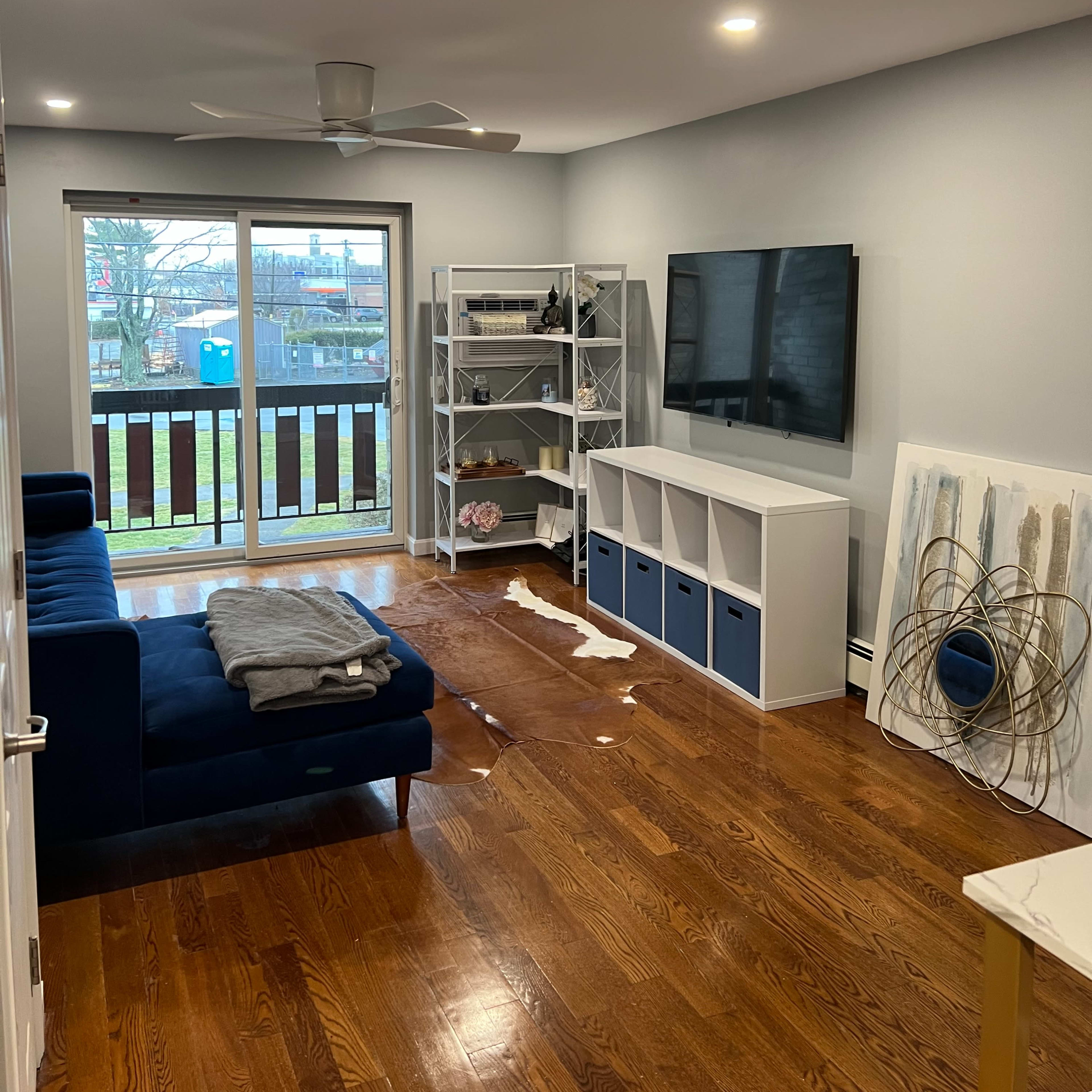 A living room features a dark blue sofa, a TV mounted on the wall, and a bookshelf filled with decorative items, next to sliding glass doors leading to a balcony.
