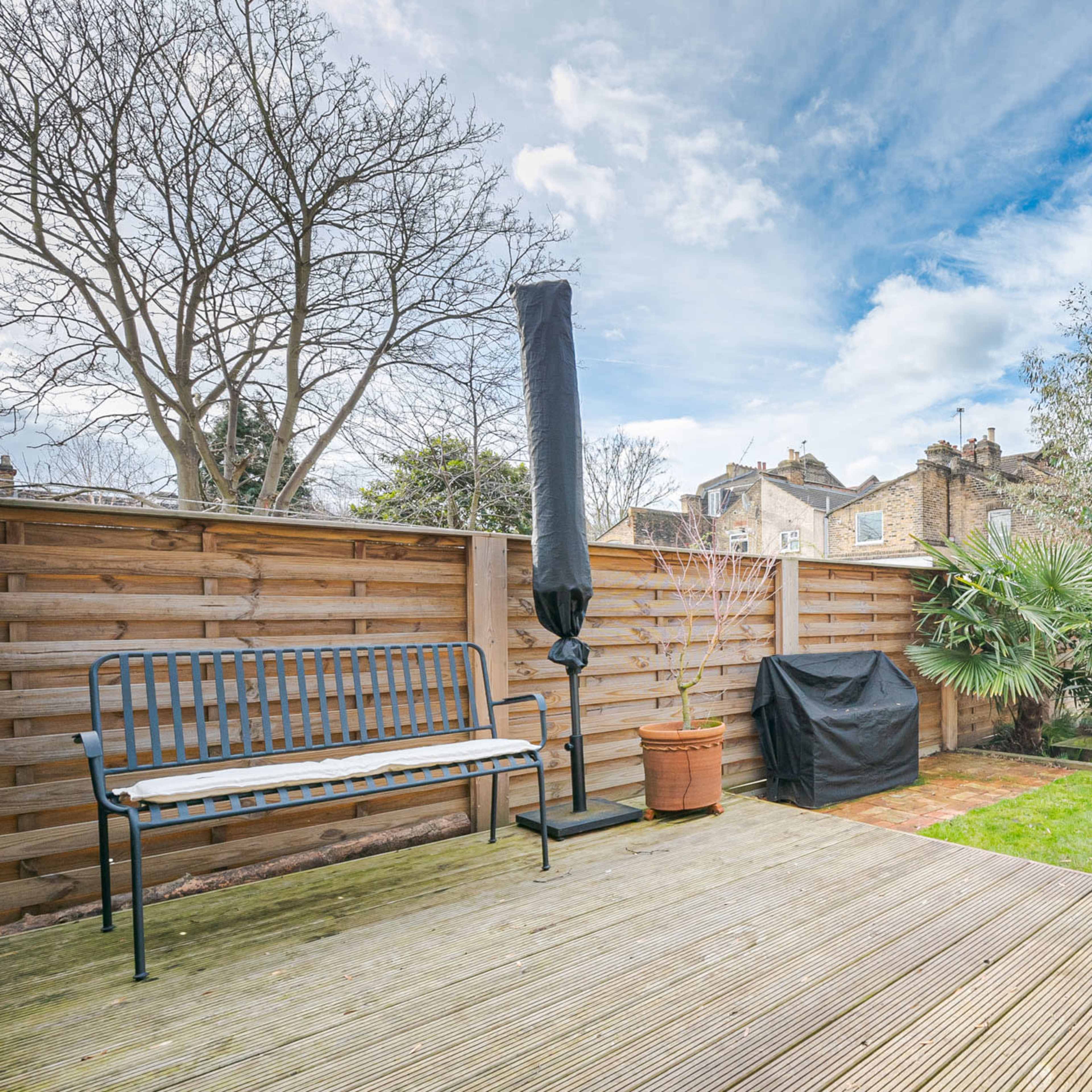 A landscaped backyard featuring a wooden deck, a bench, a potted plant, and a covered outdoor structure, with trees and shrubs in the background.