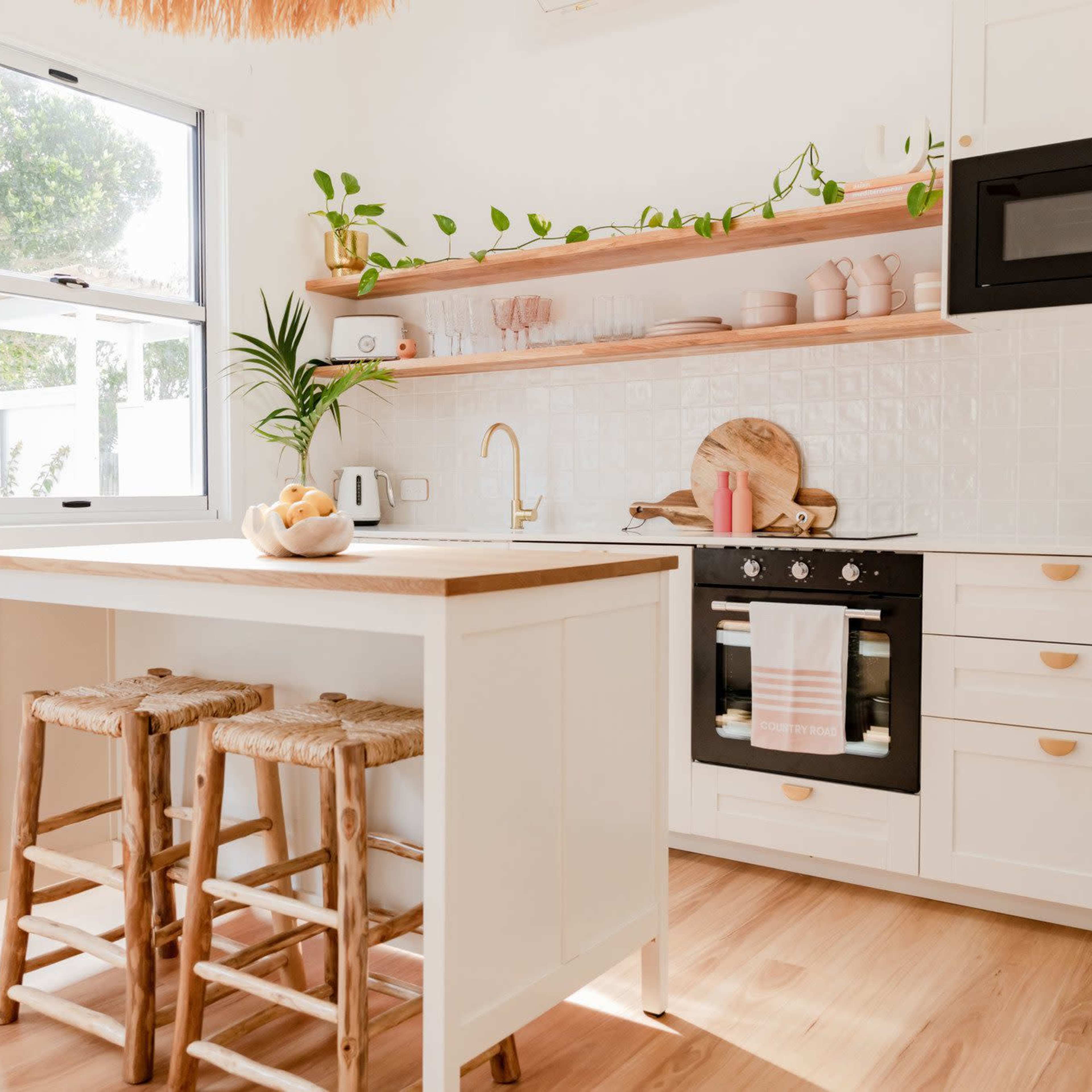 A bright kitchen features open shelving with pink dishware, a wooden island with bar stools, and modern appliances against a white backdrop.