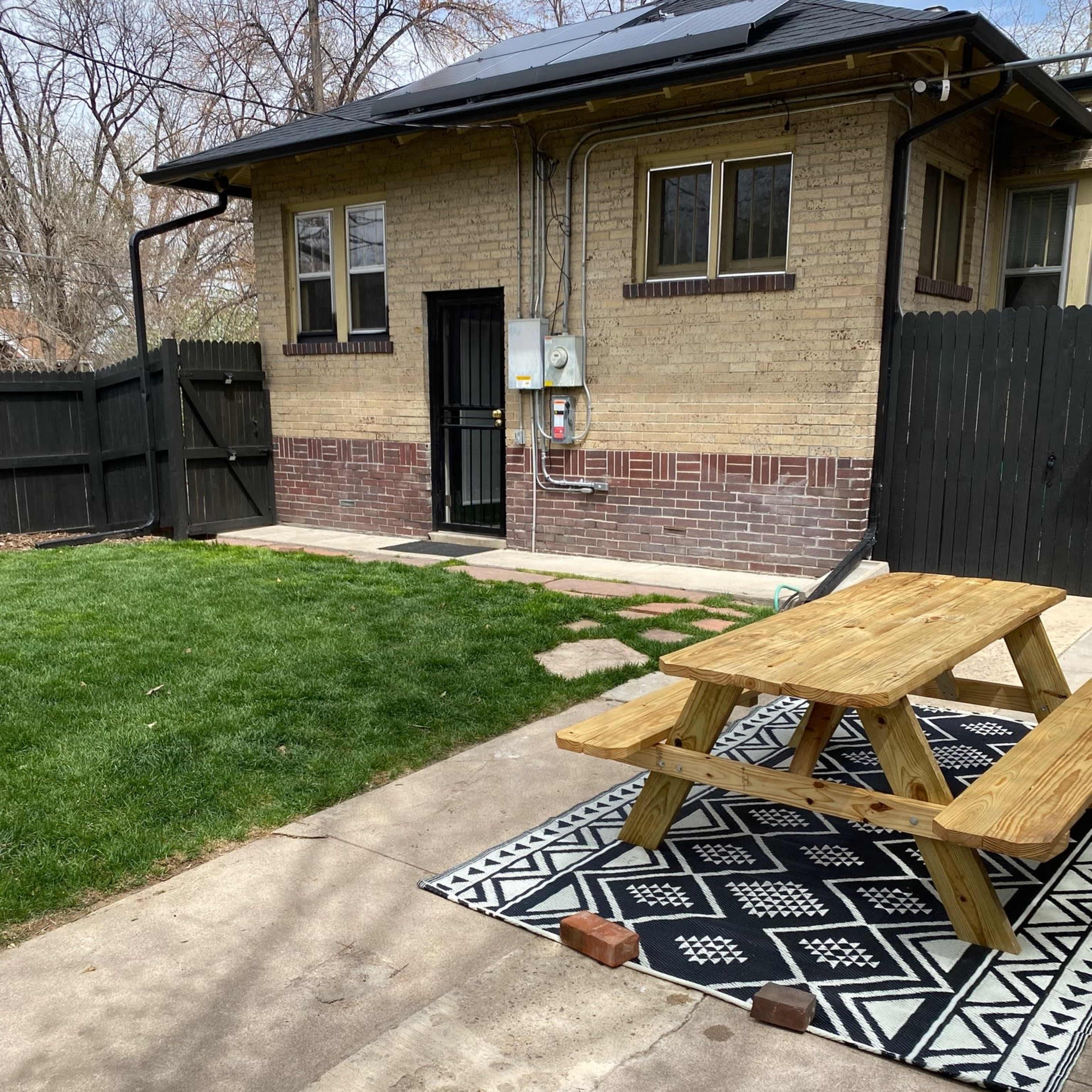 A small backyard area featuring a picnic table on a patterned rug, a patch of grass, and an entrance to a brick building with solar panels on the roof.