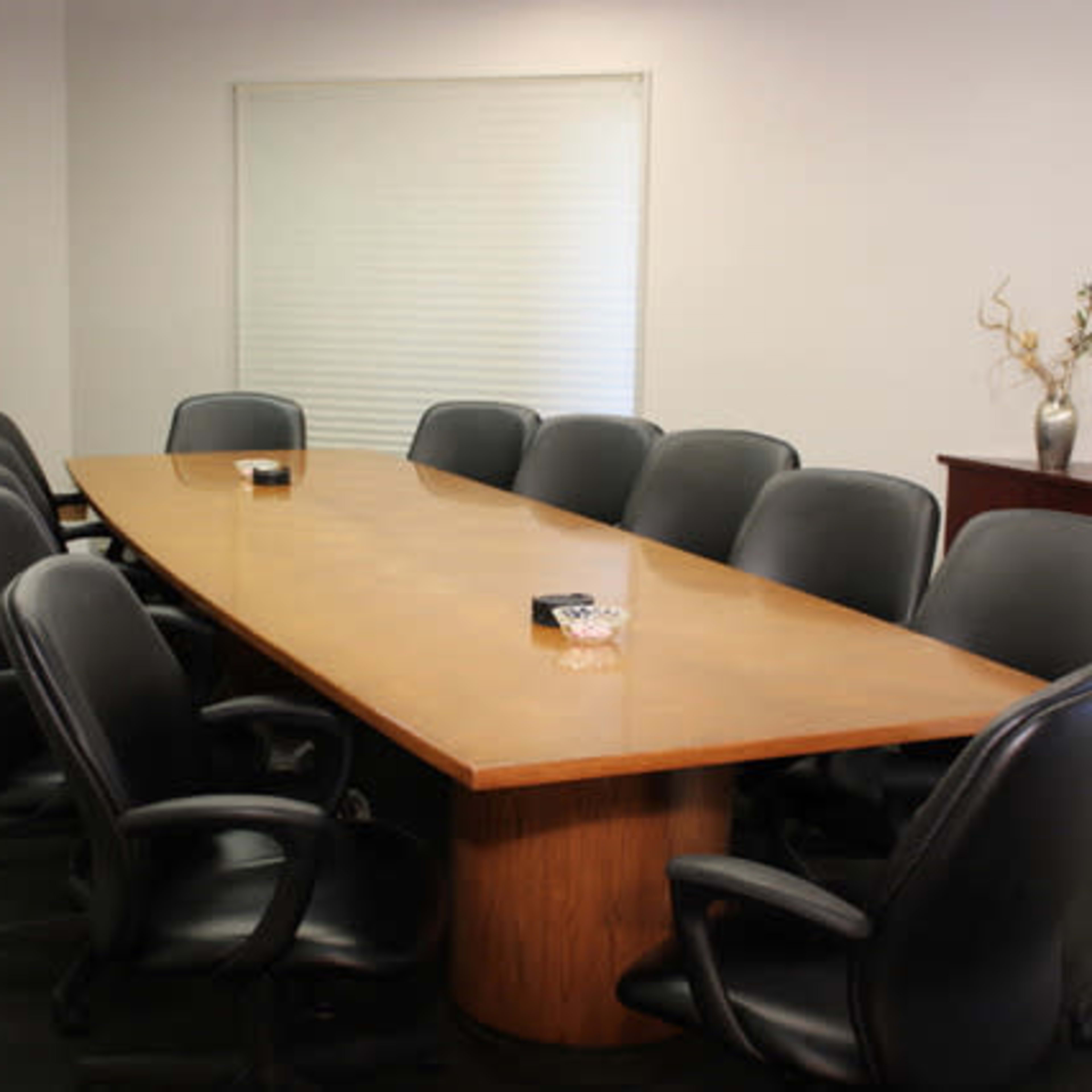 A conference room features a long wooden table surrounded by black ergonomic chairs, with a window and television in the background.