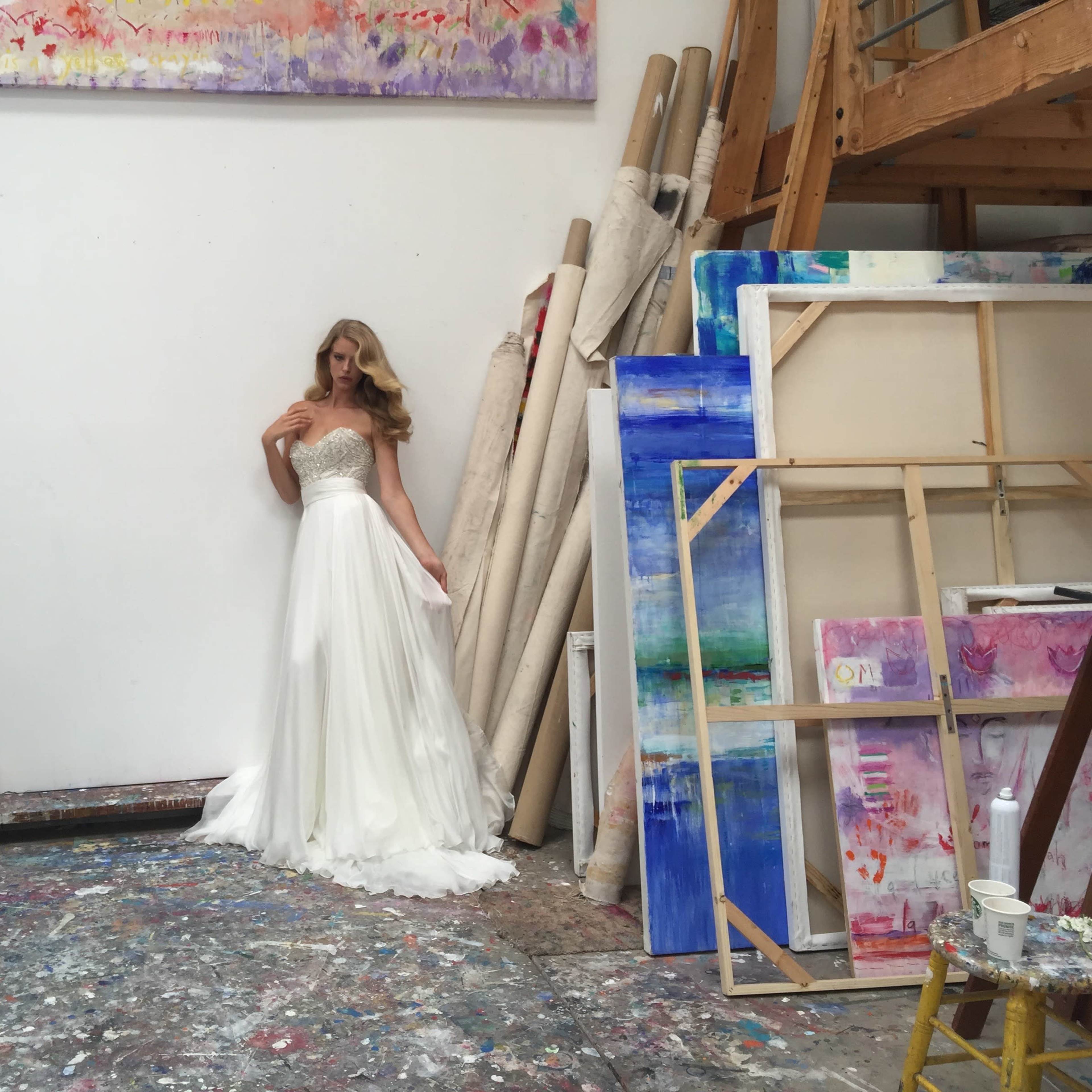 A spacious artist's studio with large skylights, colorful paintings on the walls, and scattered art supplies on the floor.