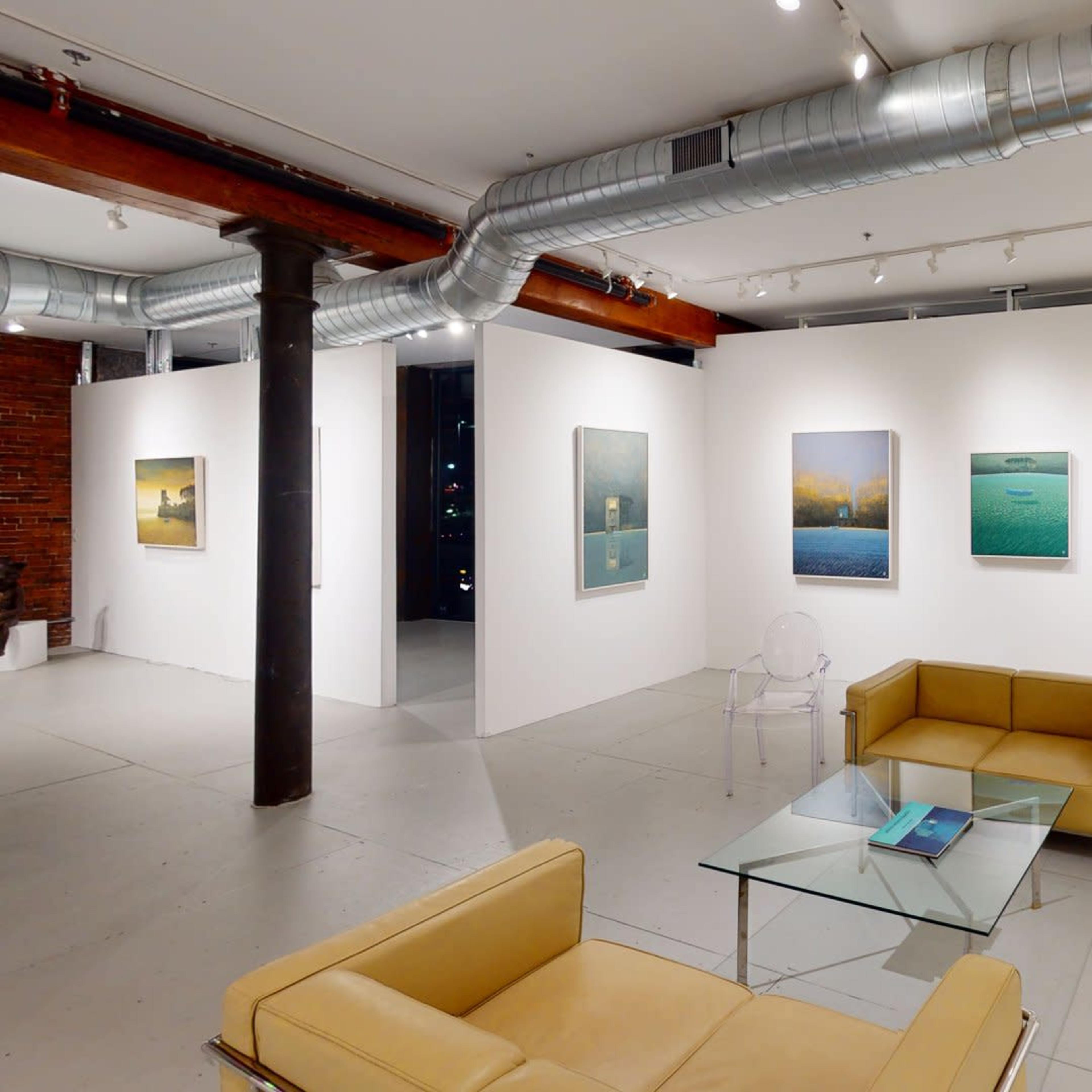 A modern art gallery with white walls displaying framed paintings, featuring a seating area with a glass table and yellow couches.