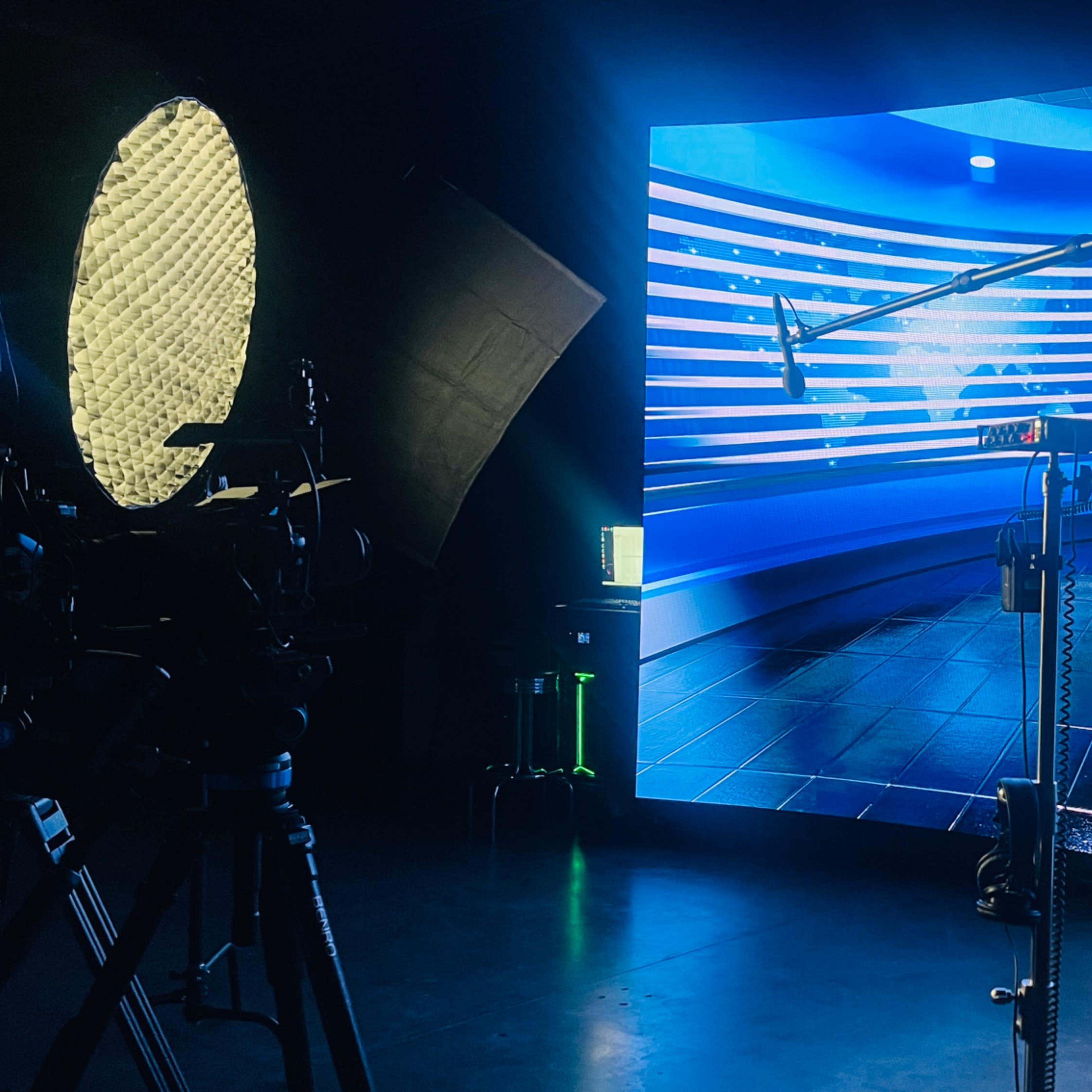 A television studio setup features a large LED screen displaying a stylized background, with cameras and lighting equipment positioned in front.