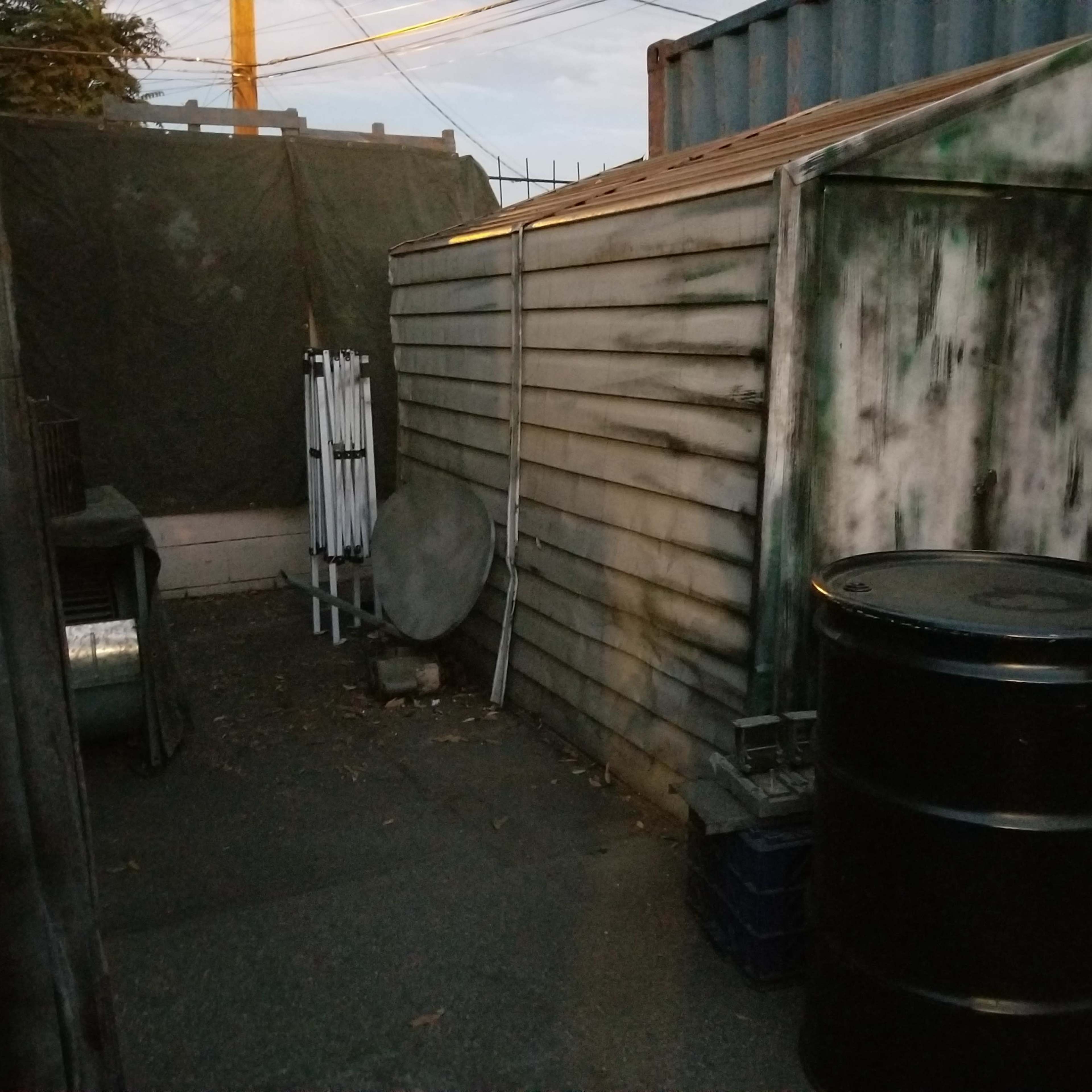 A narrow alleyway with a metal shed, a black barrel, and various equipment in a dimly lit setting.