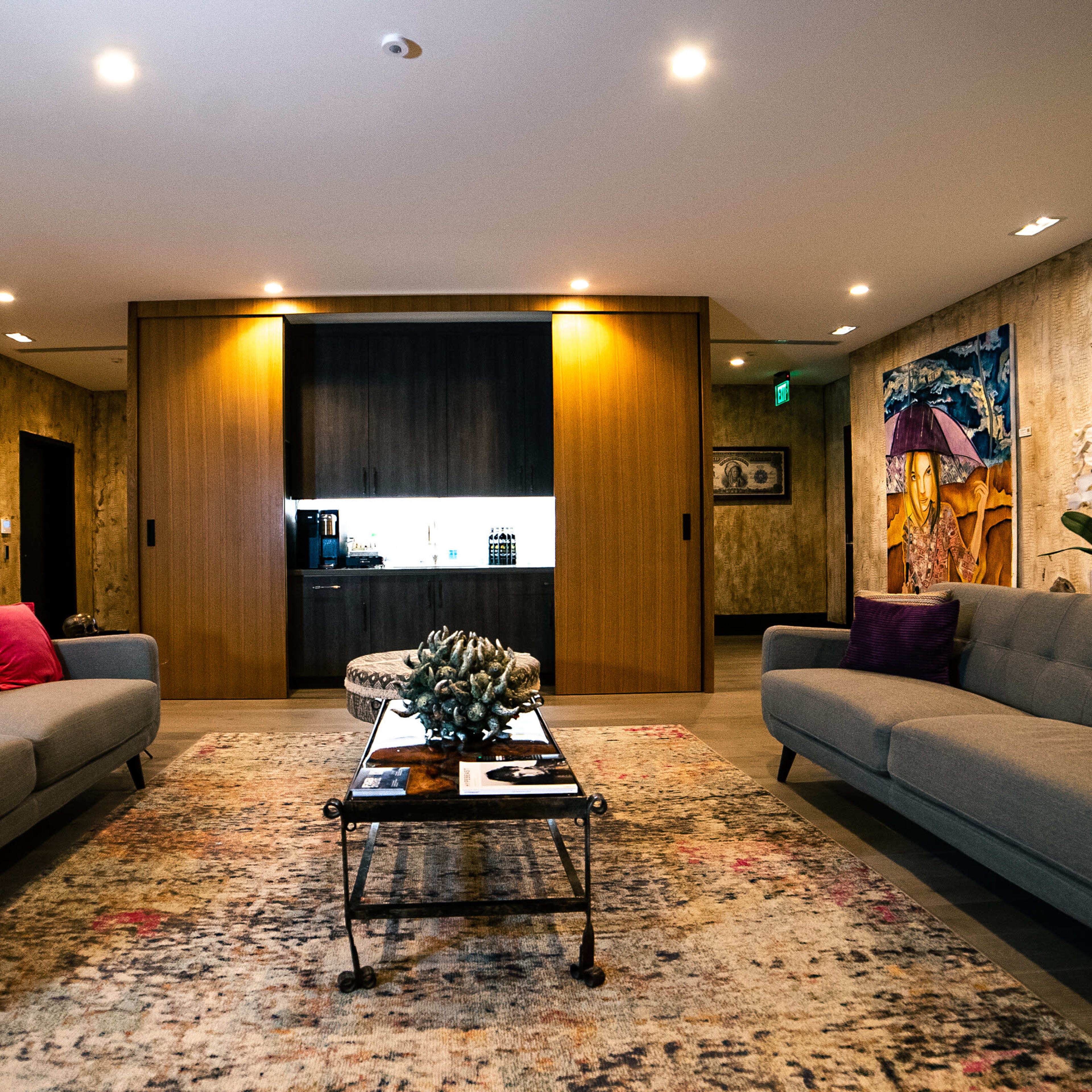 A well-lit living room with two sofas facing a coffee table, surrounded by artwork on the walls and a doorway leading to a kitchen area in the background.