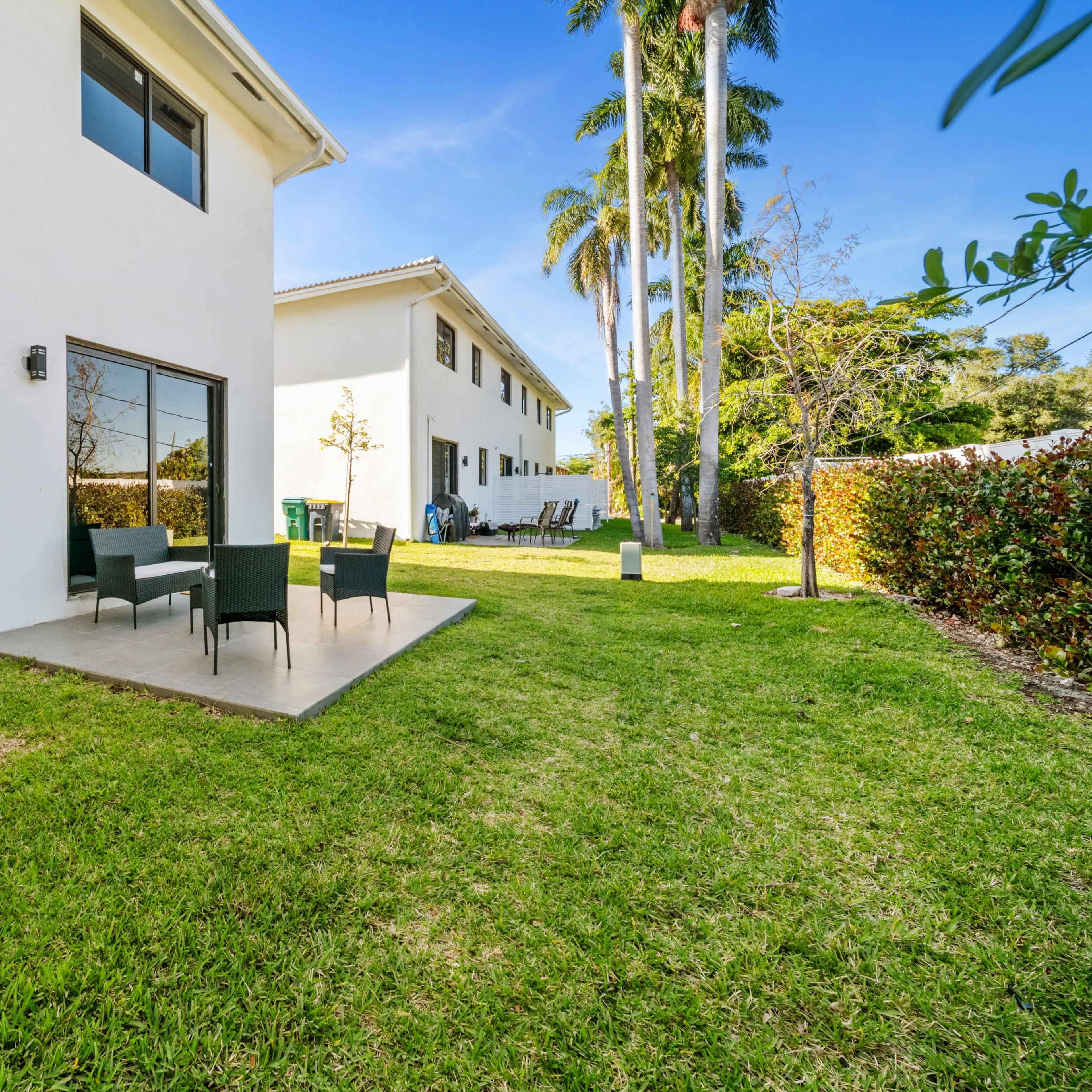 A grassy backyard with a patio featuring four outdoor chairs and two white buildings in the background.