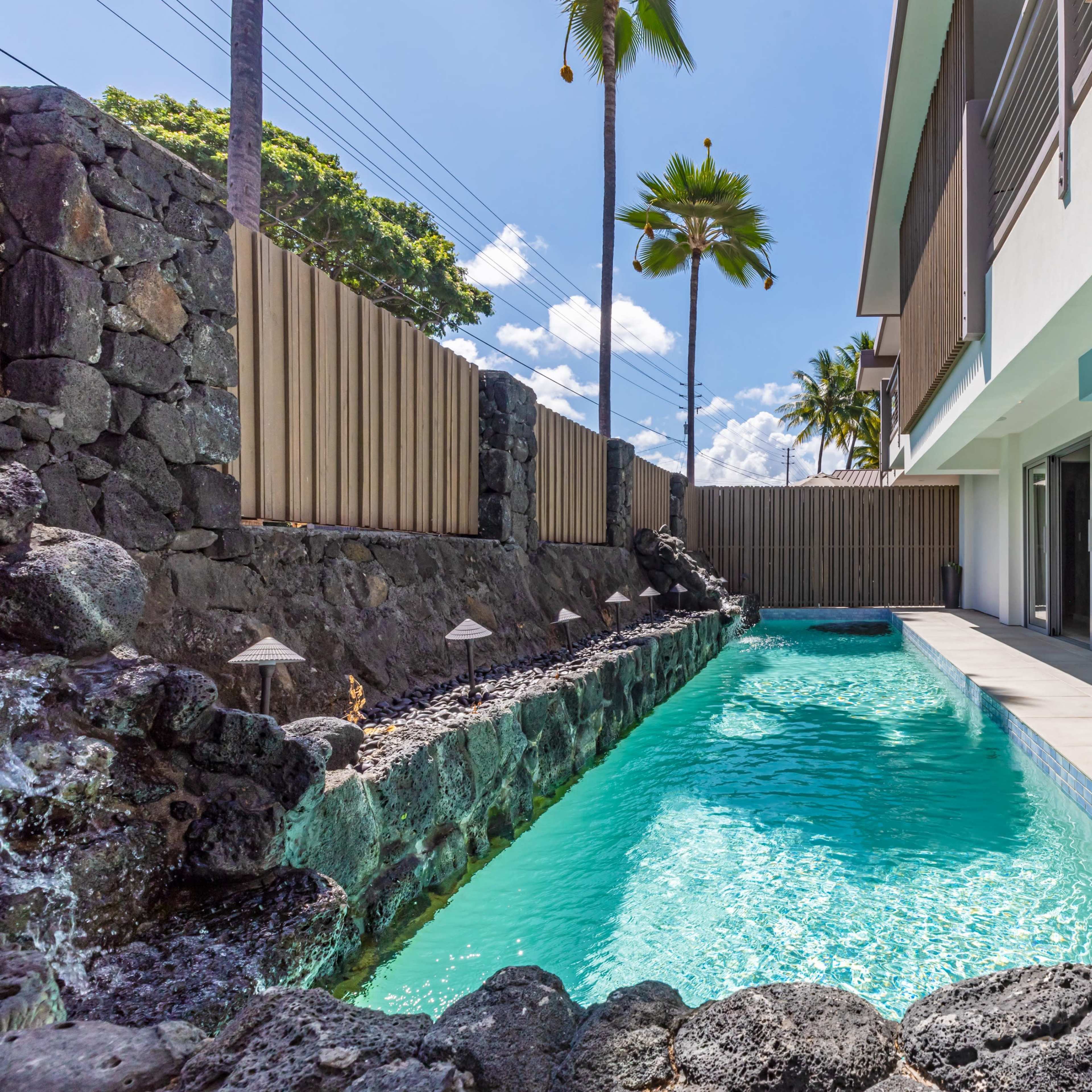 A modern swimming pool surrounded by a stone wall and palm trees, with a two-story building in the background.