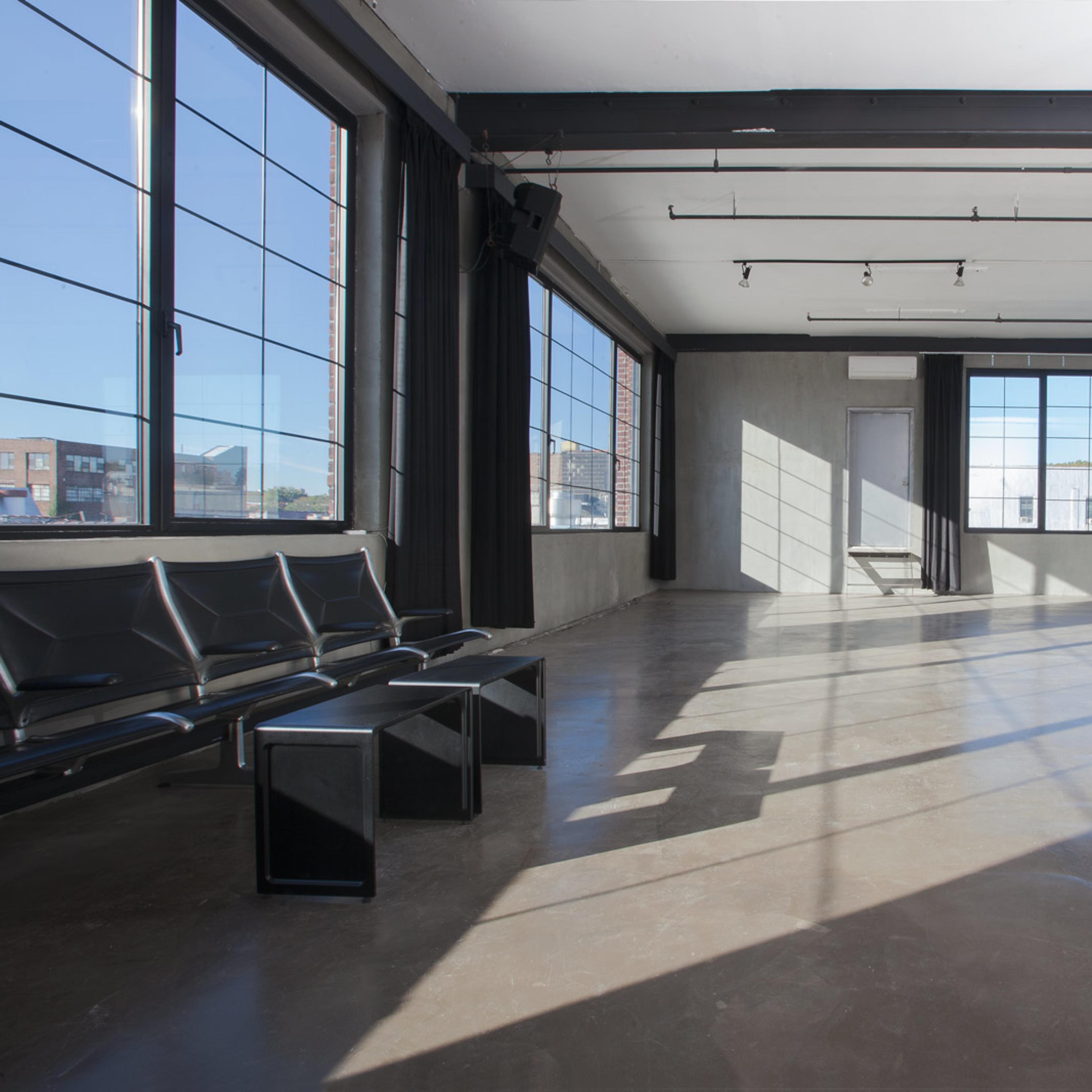 An empty industrial-style room with large windows, polished concrete flooring, and a few black chairs against one wall.