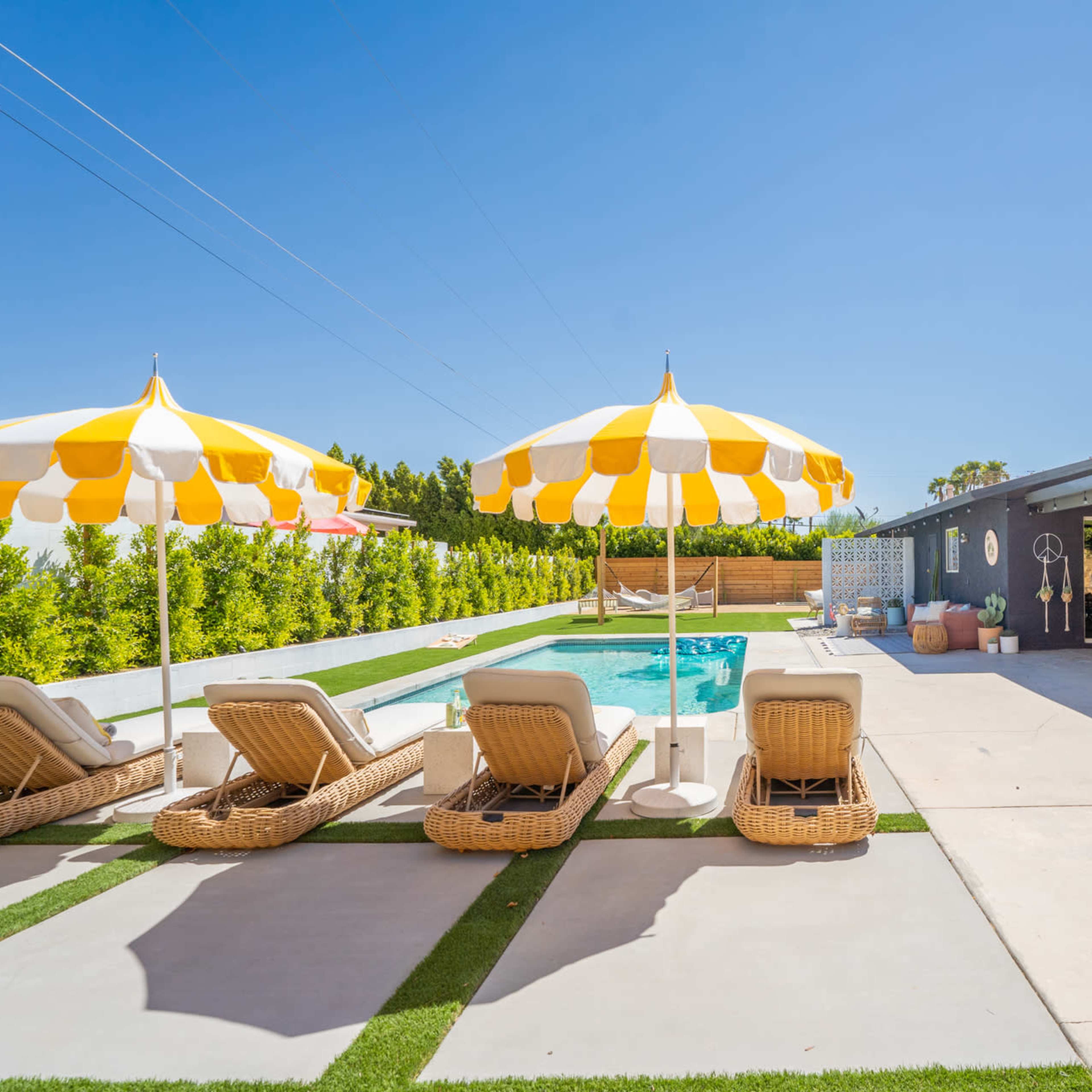 A pool area featuring lounge chairs with yellow and white striped umbrellas beside a swimming pool, surrounded by greenery and a modern home.