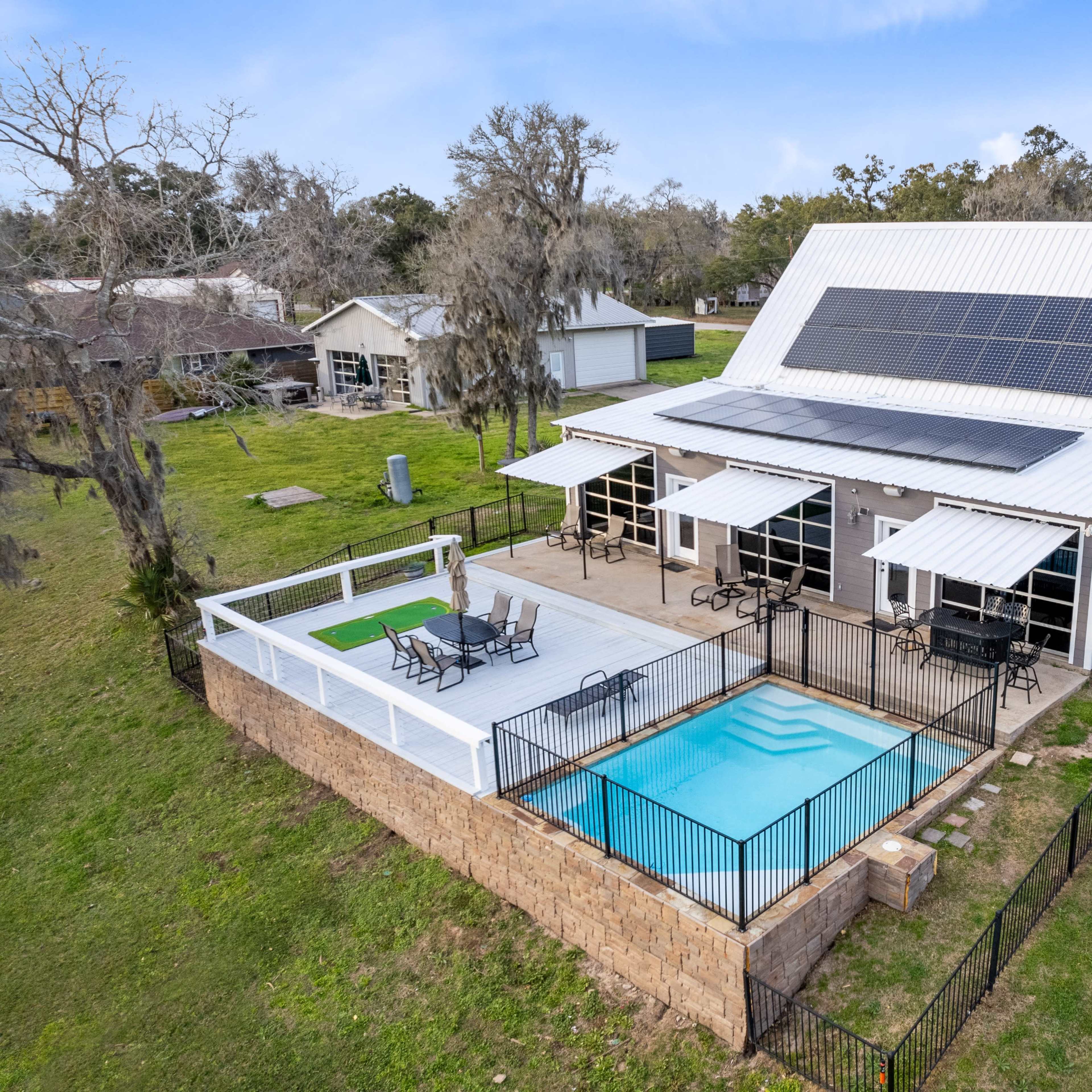 A modern house with a metal roof, solar panels, and a fenced swimming pool surrounded by a wooden deck and patio furniture.