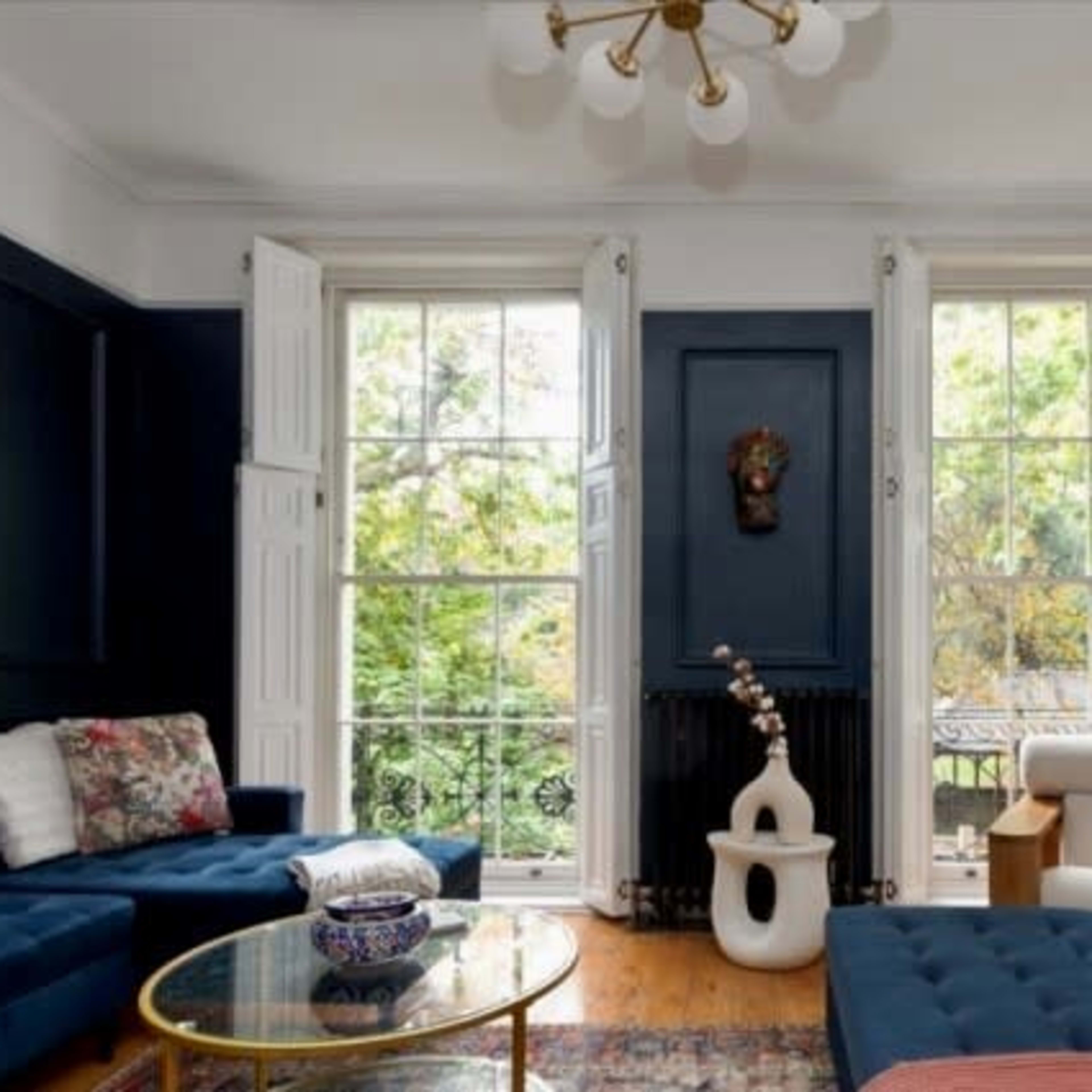 A living room featuring dark blue walls, large windows with white shutters, and a mix of blue and white furniture arrangements.
