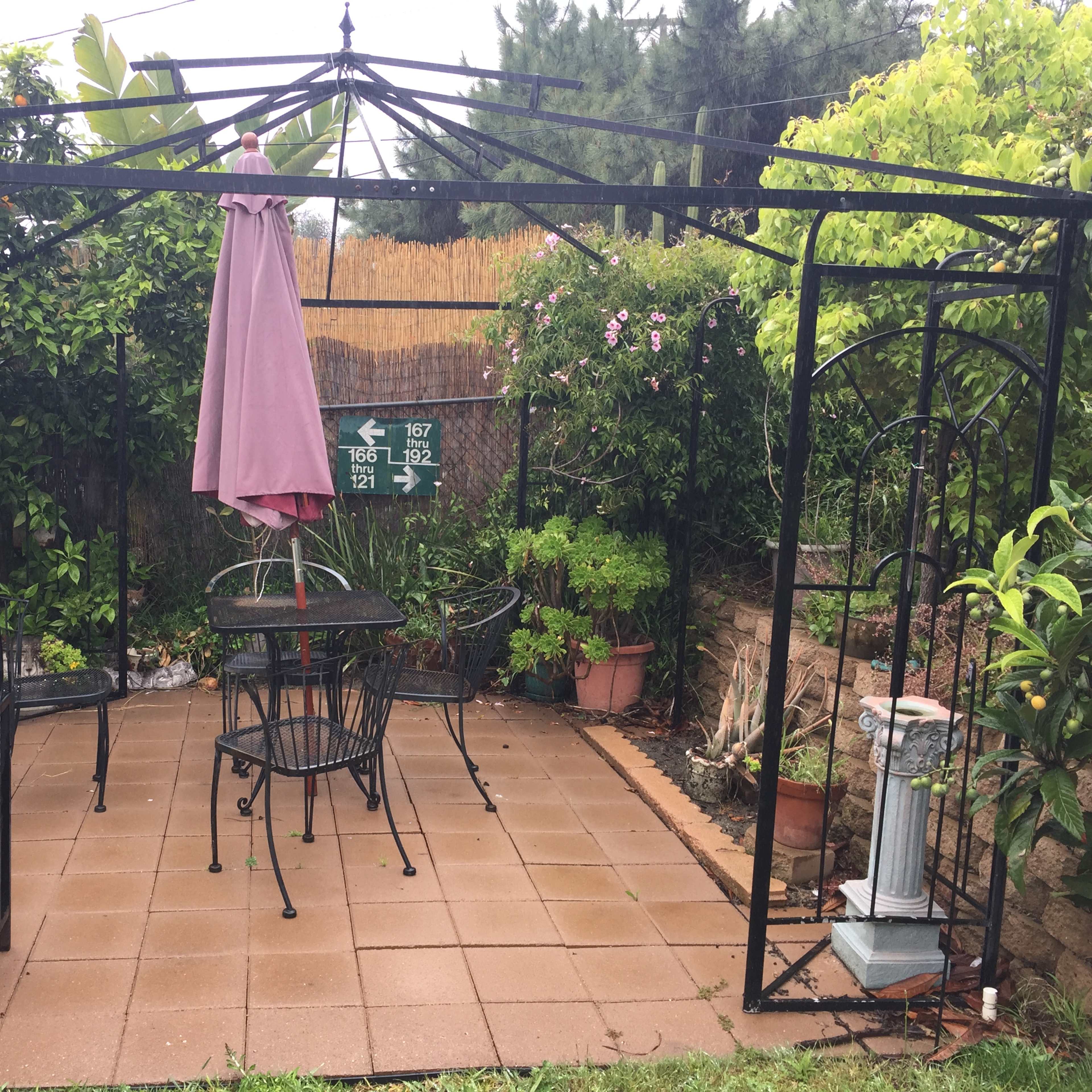 A patio area features a black metal frame with a pink umbrella, surrounded by potted plants and two black chairs and a table on tiled flooring.