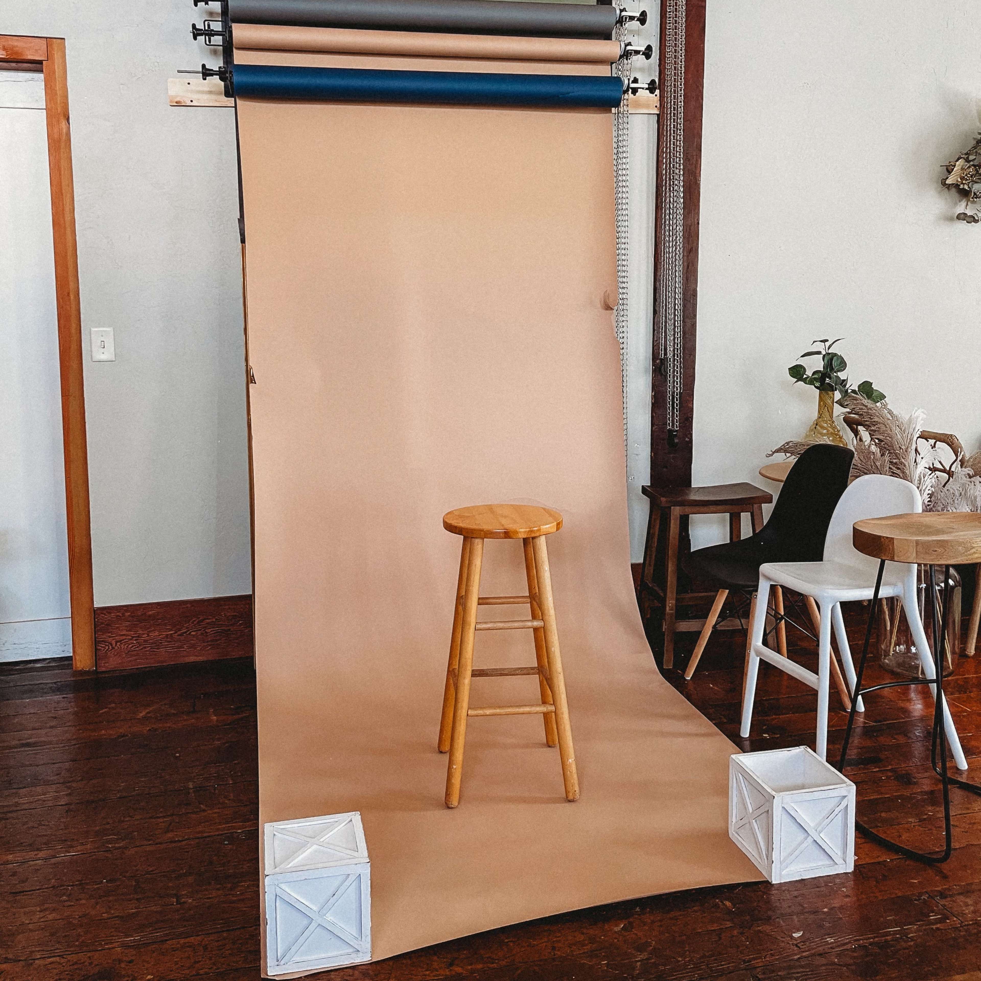 A photo studio setup with a wooden stool positioned in front of a tan backdrop and rolls of colored paper hanging above.