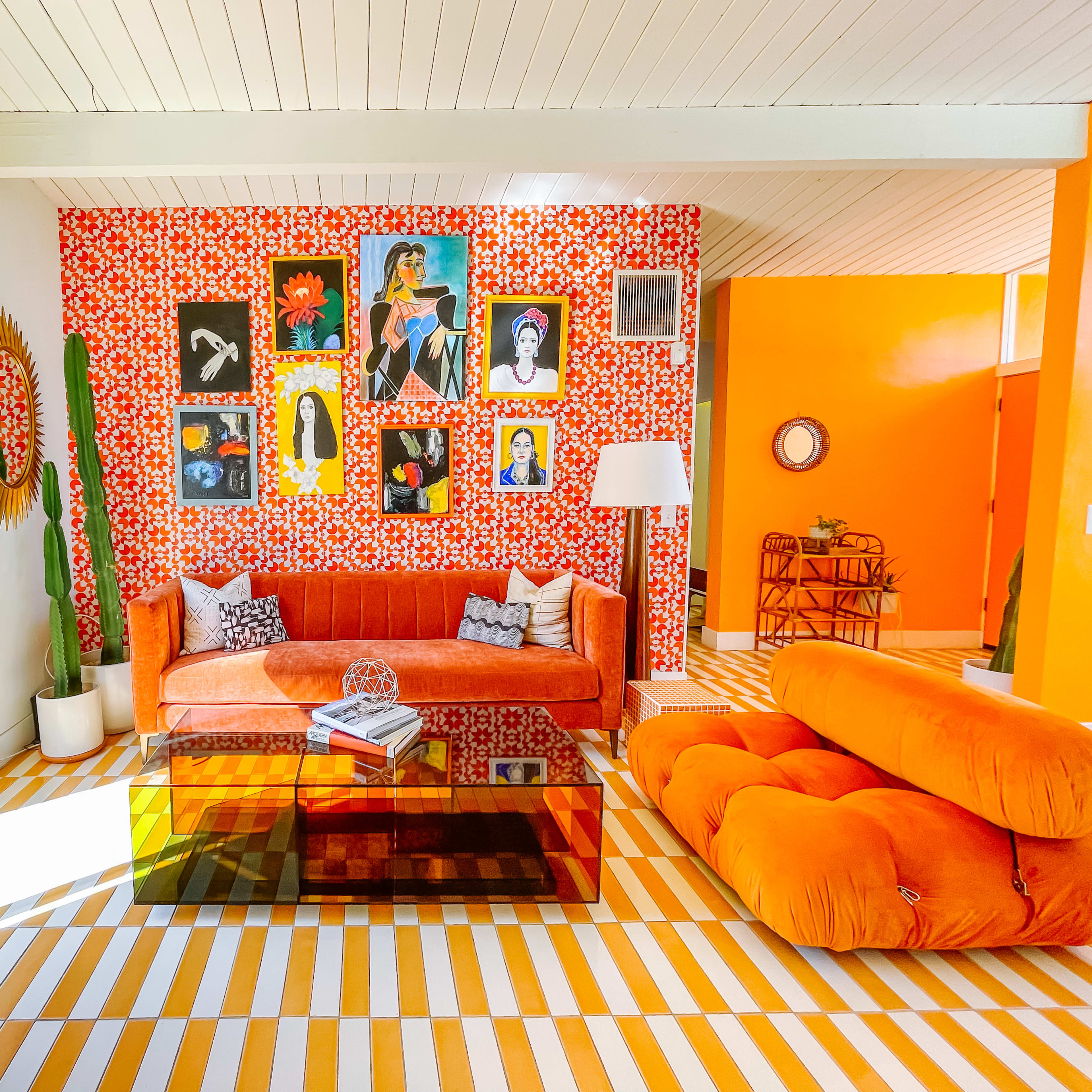 The living room features a wall adorned with various framed artworks, an orange velvet sofa, and a glass coffee table on striped flooring.