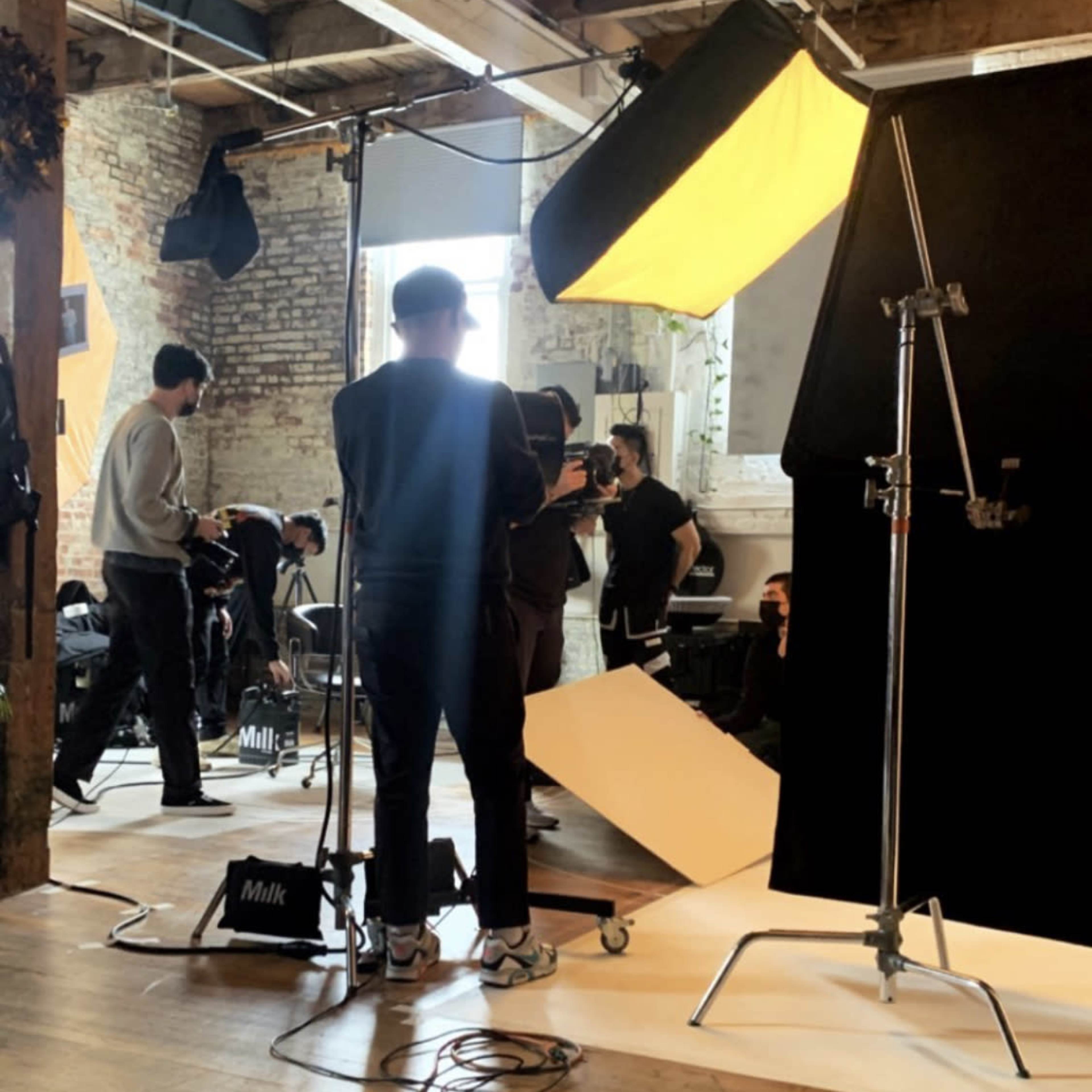 A film crew sets up equipment in a studio with wooden floors and exposed brick walls for a shoot.