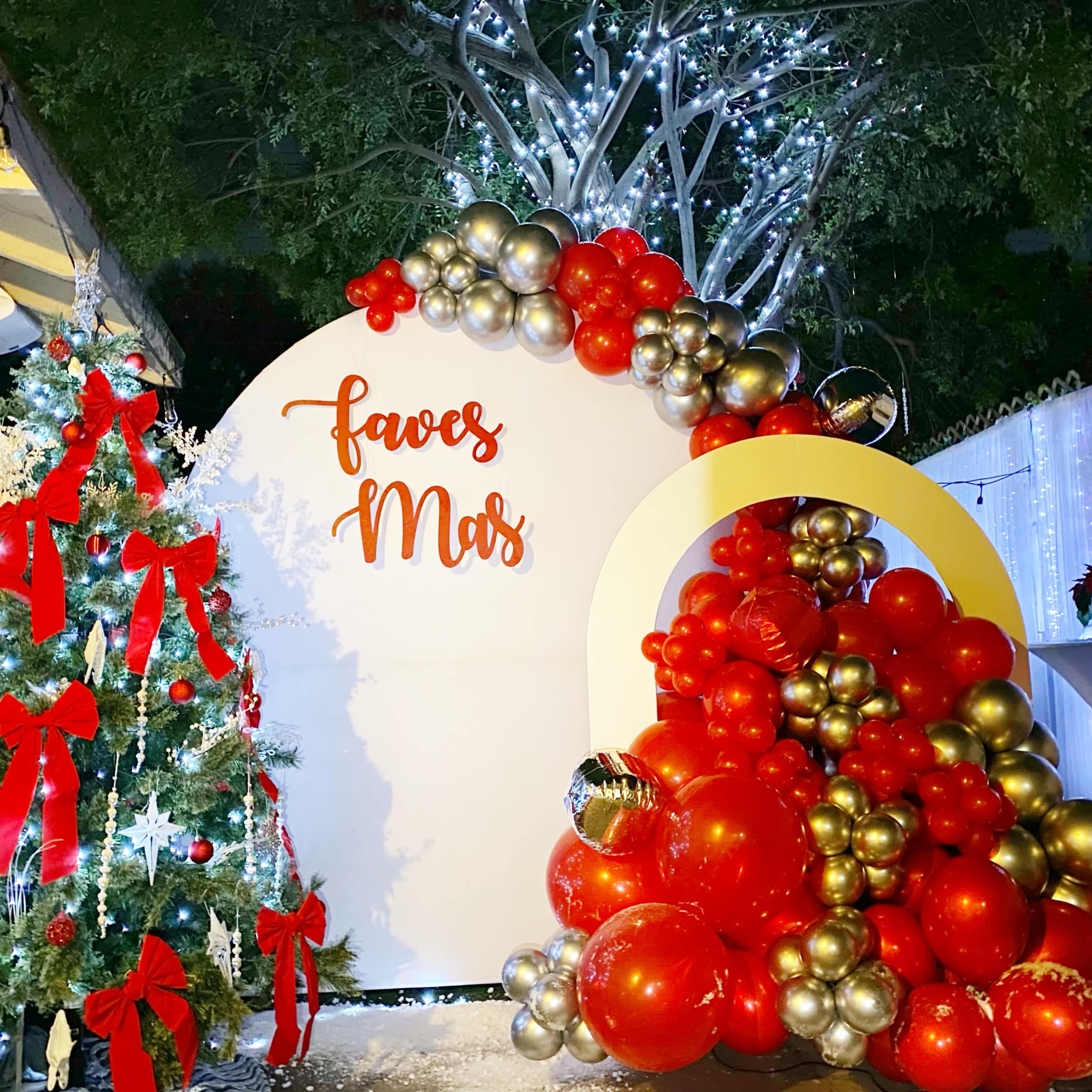 A festive display with a decorated Christmas tree on one side and a large balloon arrangement in red and gold on the other, set against a backdrop that says "faves Mar."