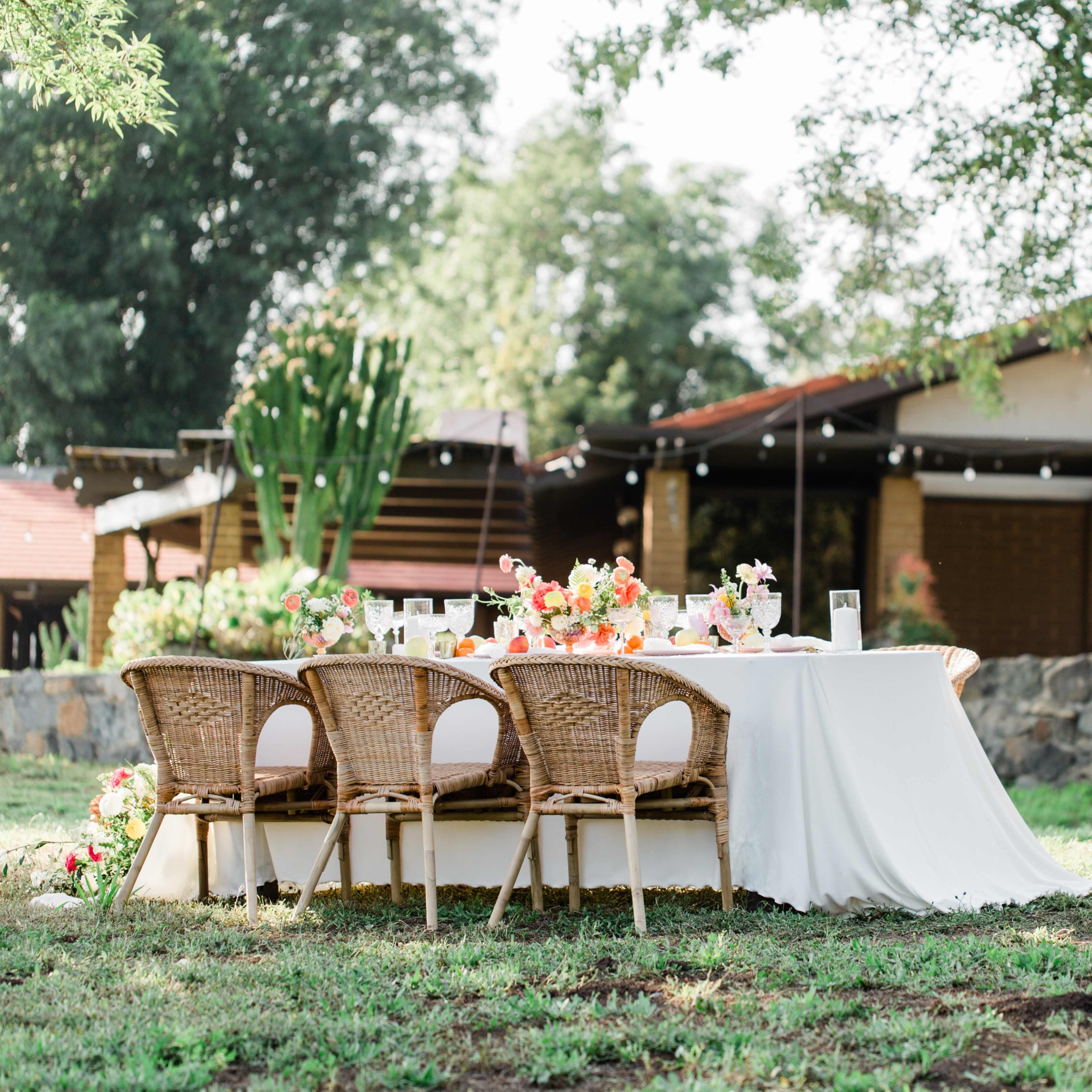 A beautifully set outdoor dining table with wicker chairs is positioned on grass in front of a house surrounded by trees.