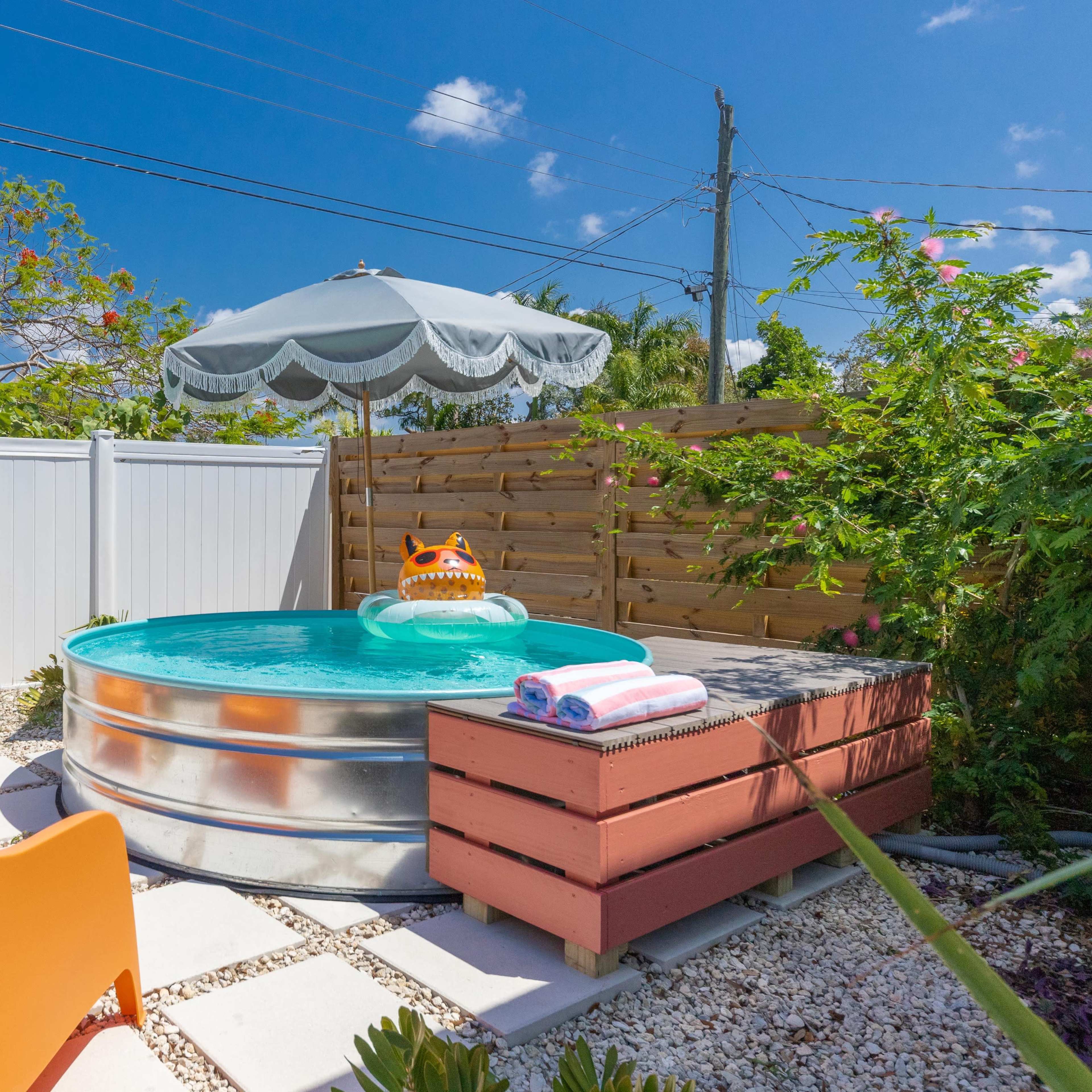 A small metal pool surrounded by stone pavers, two orange chairs, a wooden box, and a shaded umbrella in a fenced outdoor space.