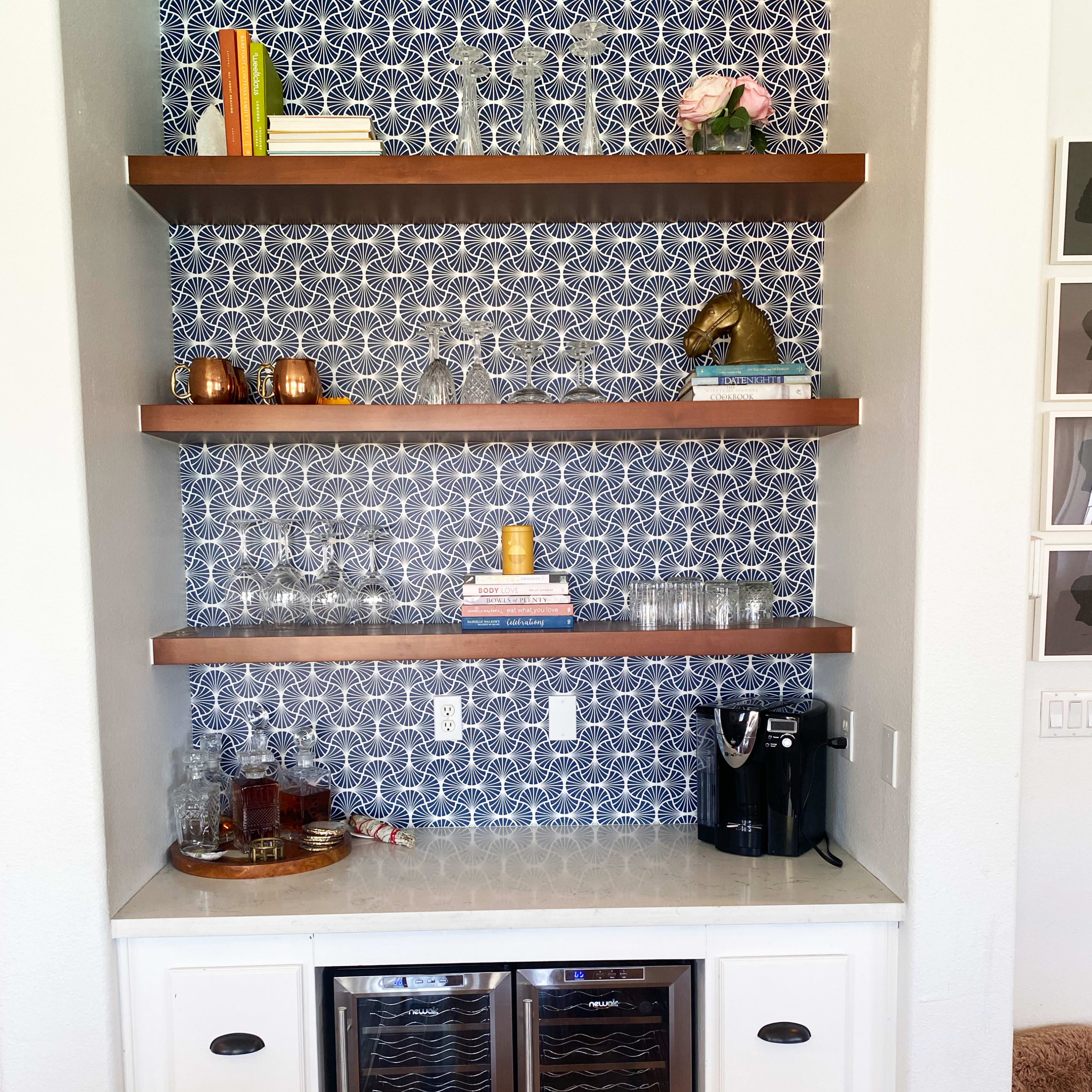 A modern kitchen nook with floating shelves displaying glassware and decor, a countertop with a coffee machine, and a wine cooler below.
