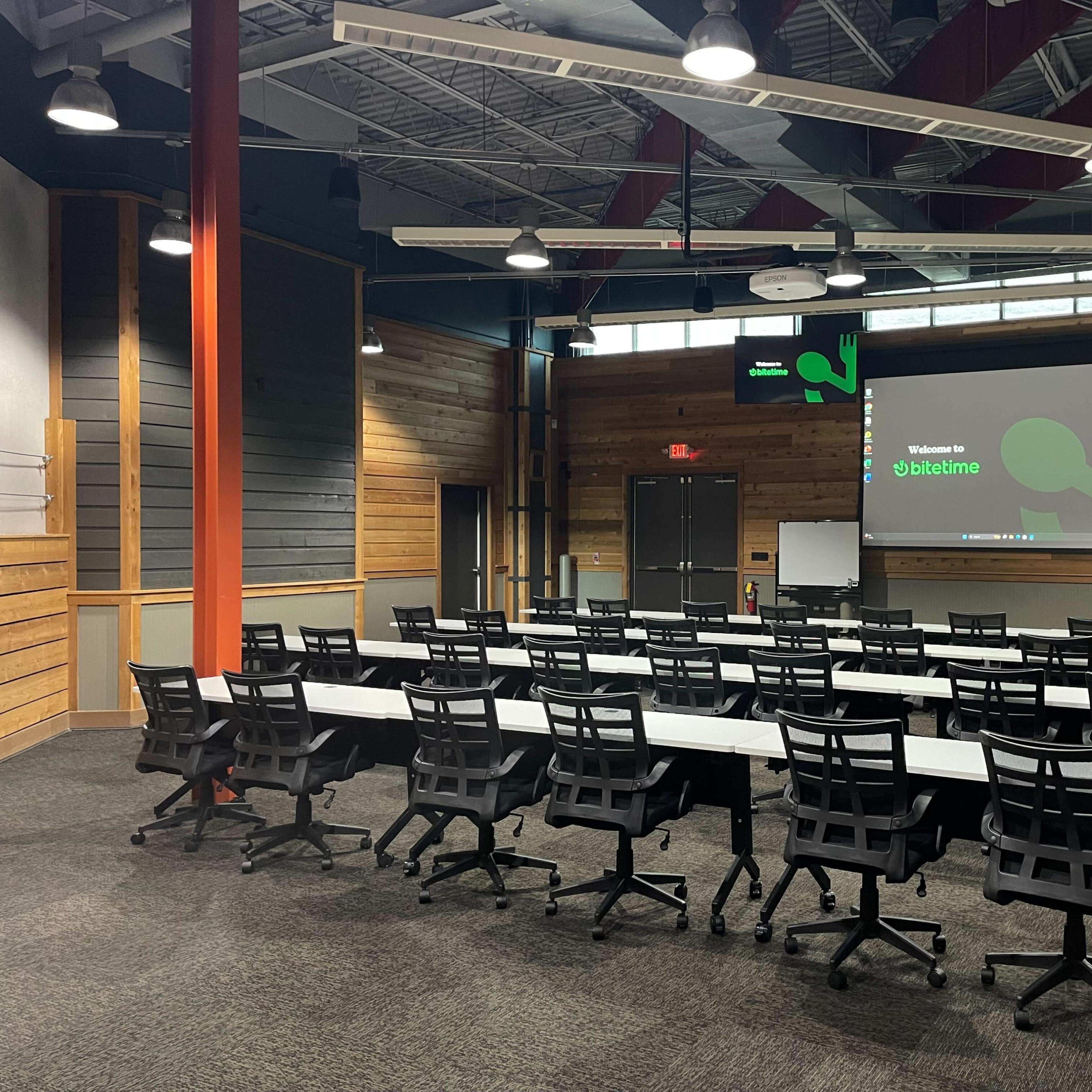 A conference room set up with rows of tables and chairs, a screen displaying a welcome message, and a modern design featuring wooden accents.