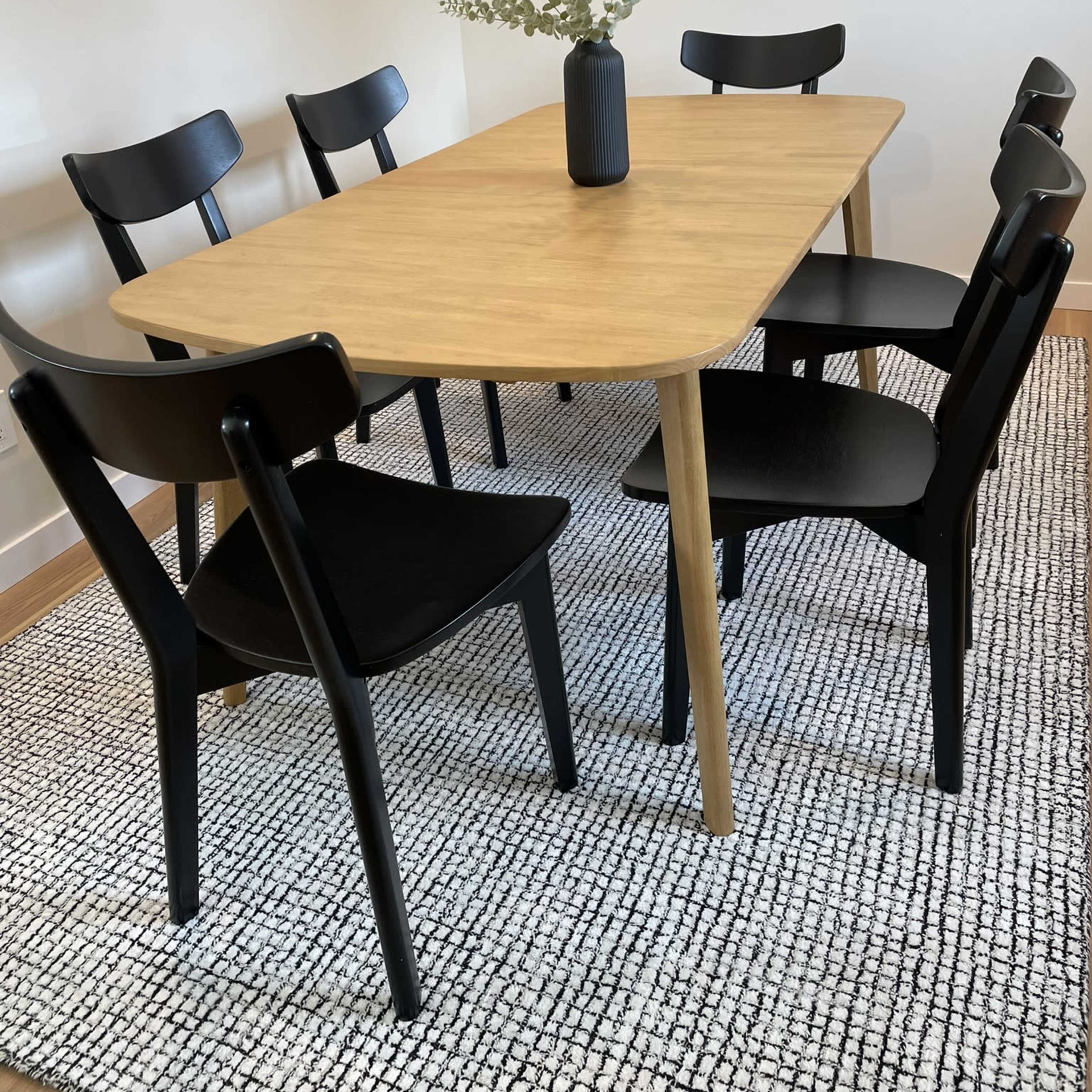 A wooden dining table surrounded by six black chairs on a textured rug with a small vase of greenery in the center.