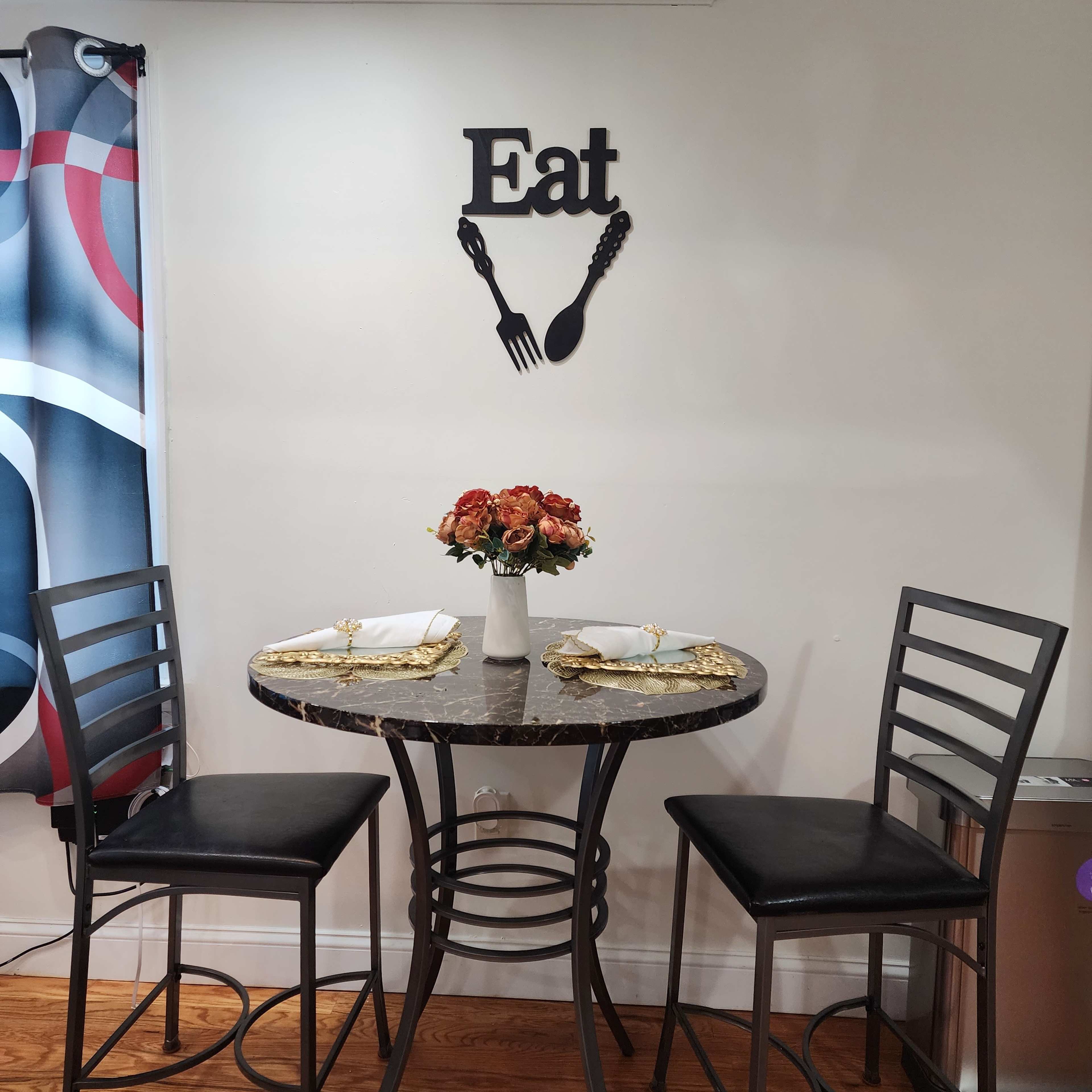 A small dining table with two black chairs is set against a wall adorned with a decorative "Eat" sign and a vase of flowers on the table.