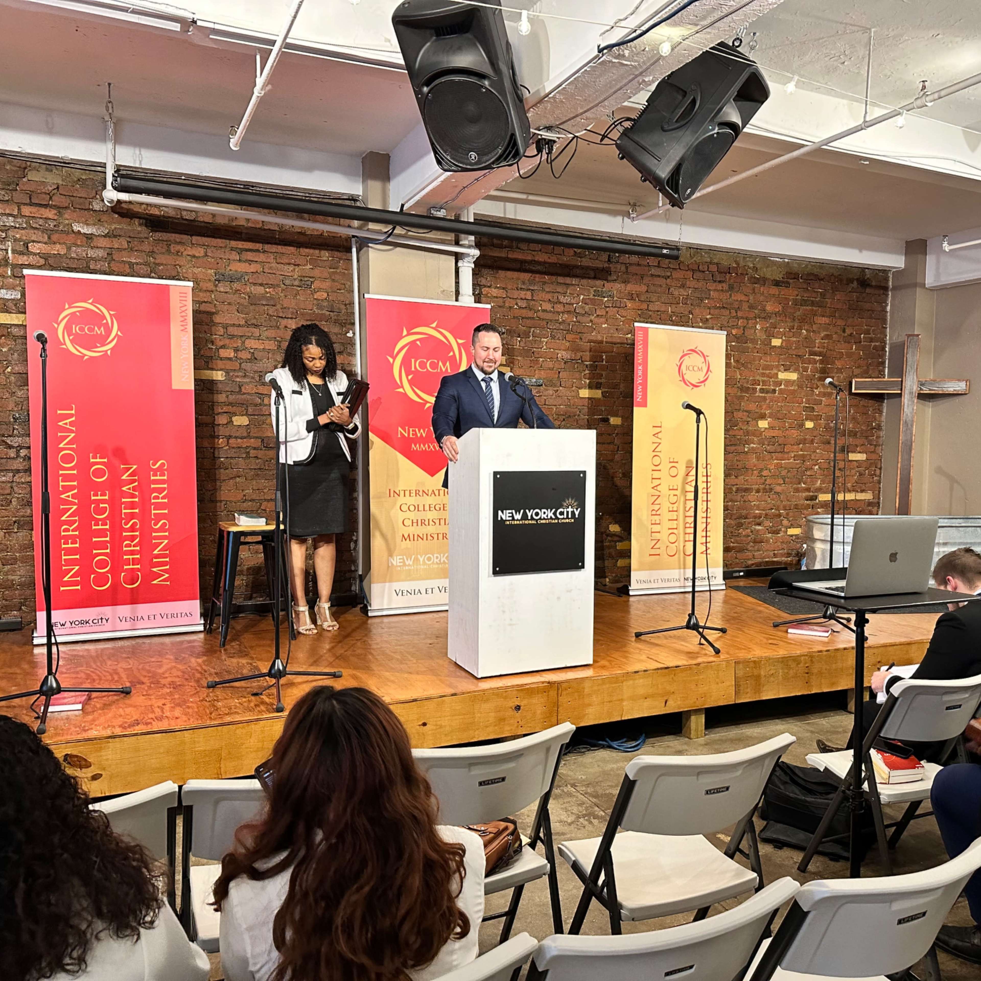 A speaker stands at a podium in front of an audience during a church event in a brick-walled venue.