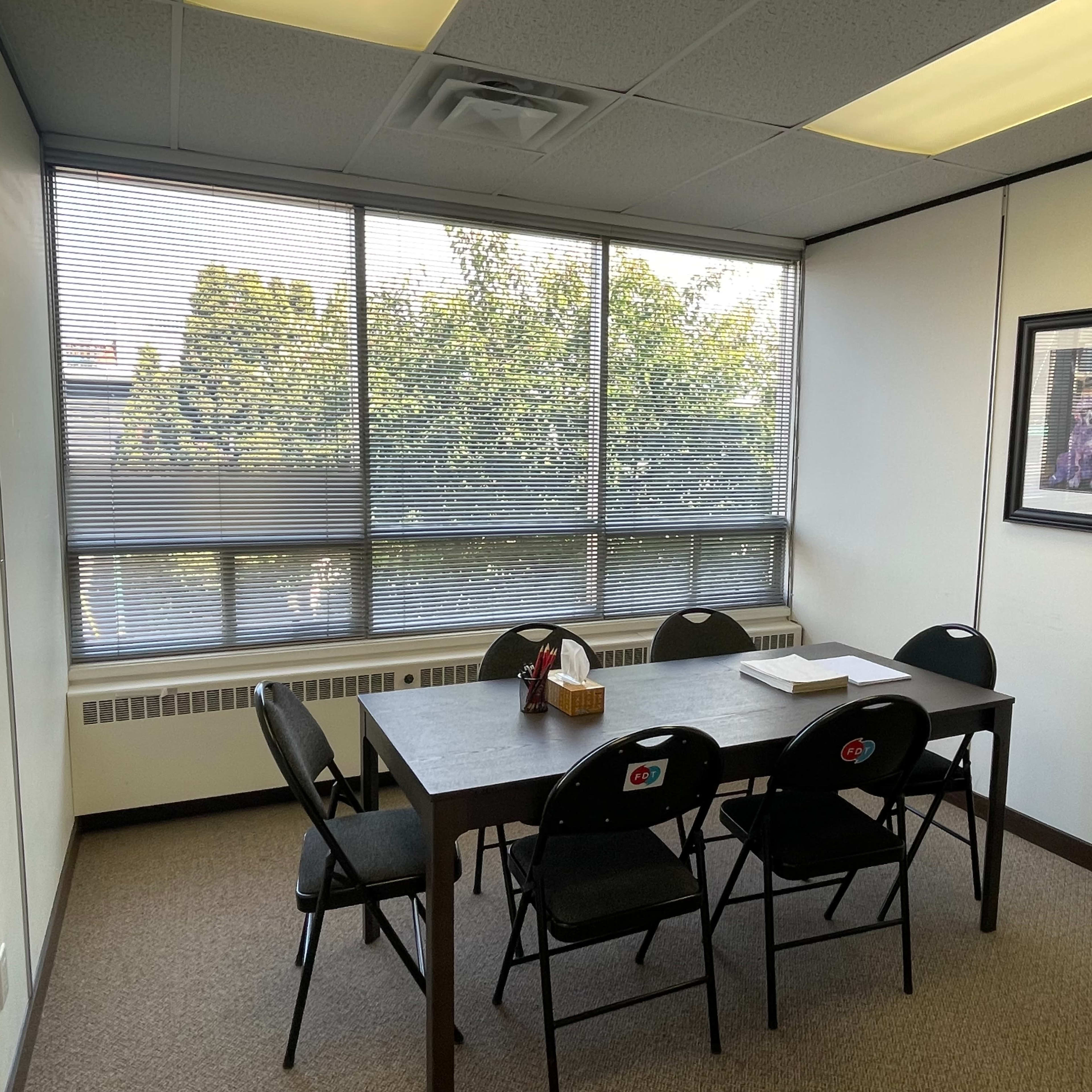 A conference room with a table surrounded by chairs, large windows providing natural light, and a framed artwork on the wall.