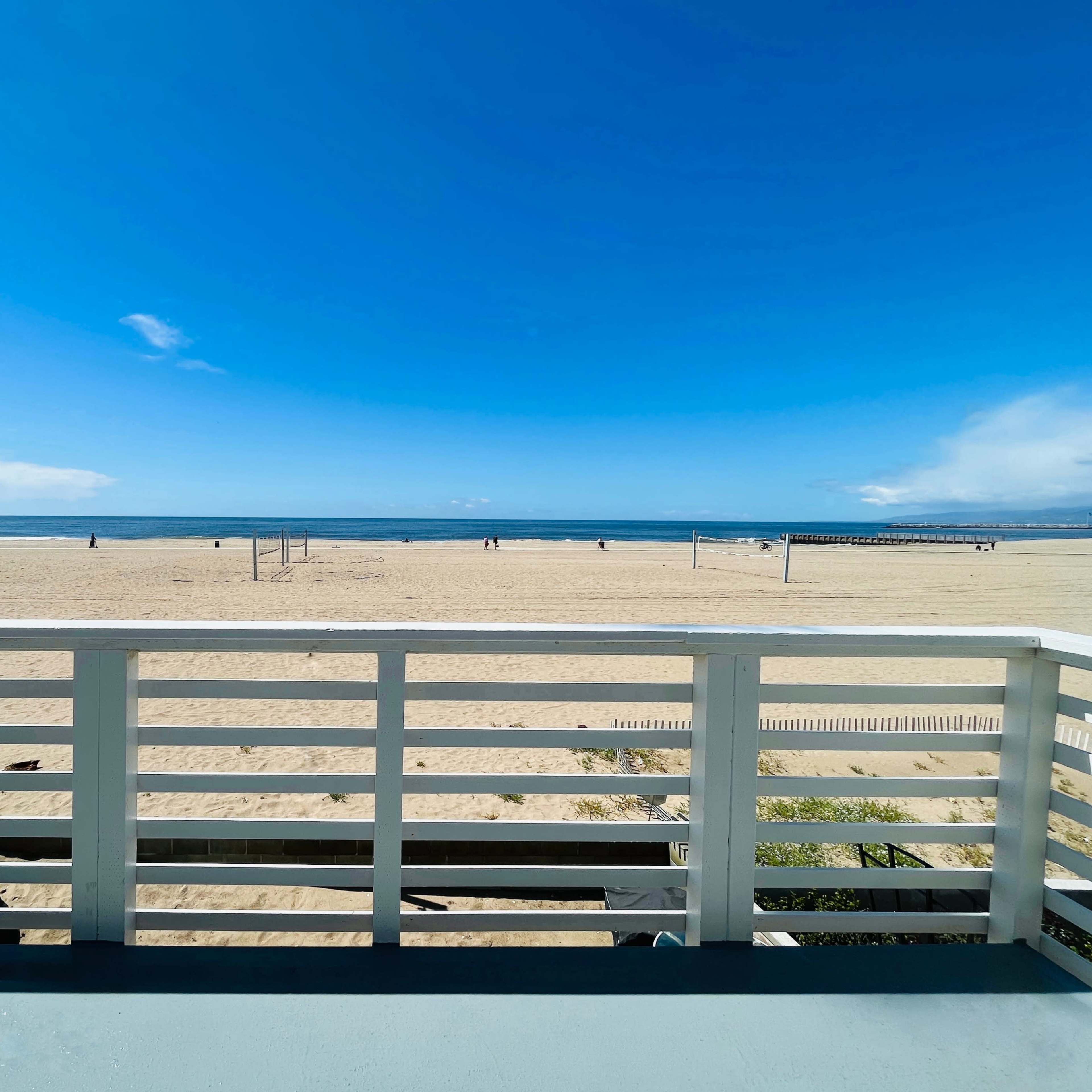 A view of a sandy beach with a blue sky and a few people in the distance, framed by a white railing and a palm tree.