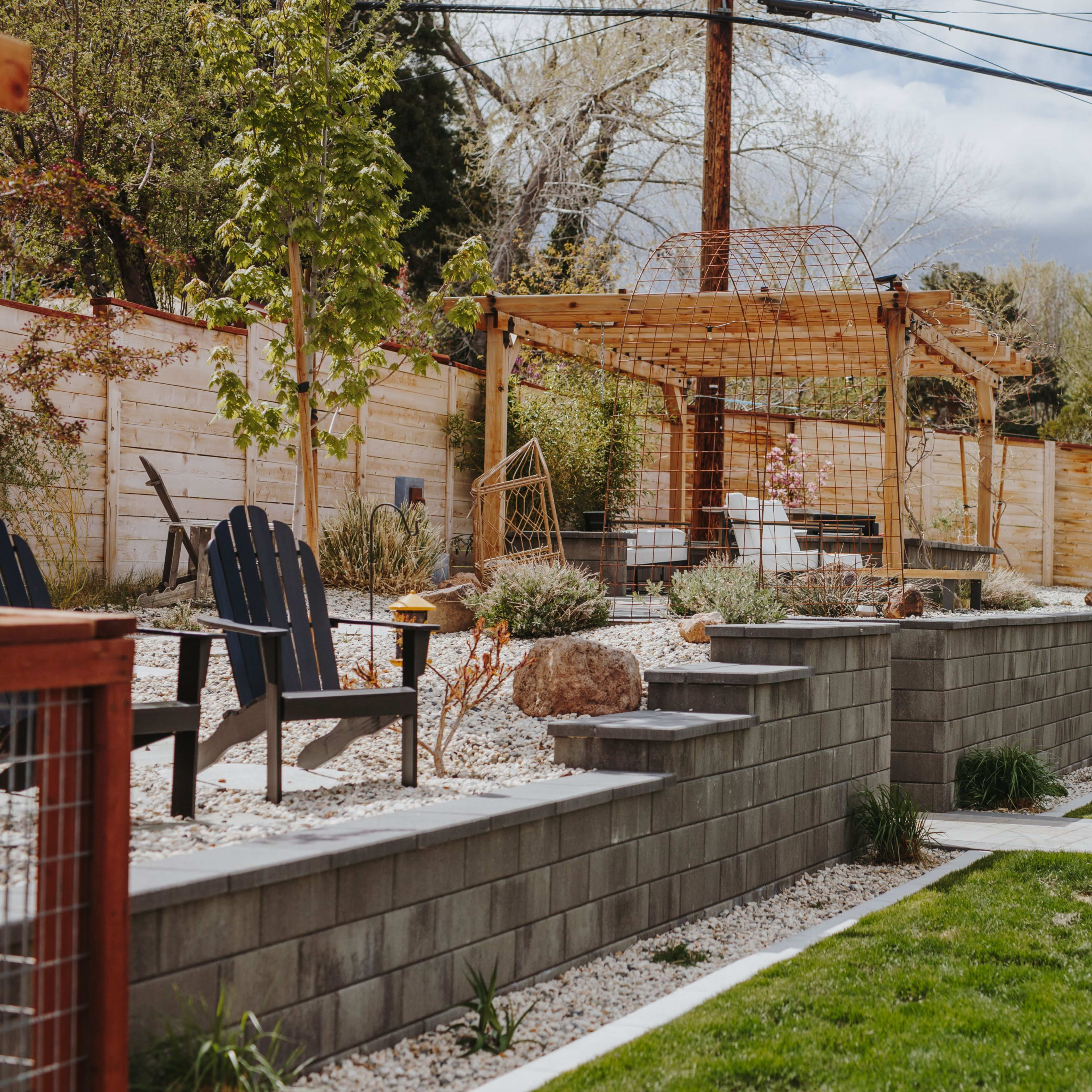 A landscaped backyard with a stone wall, gravel beds, a pergola, and wooden seating areas surrounded by greenery.