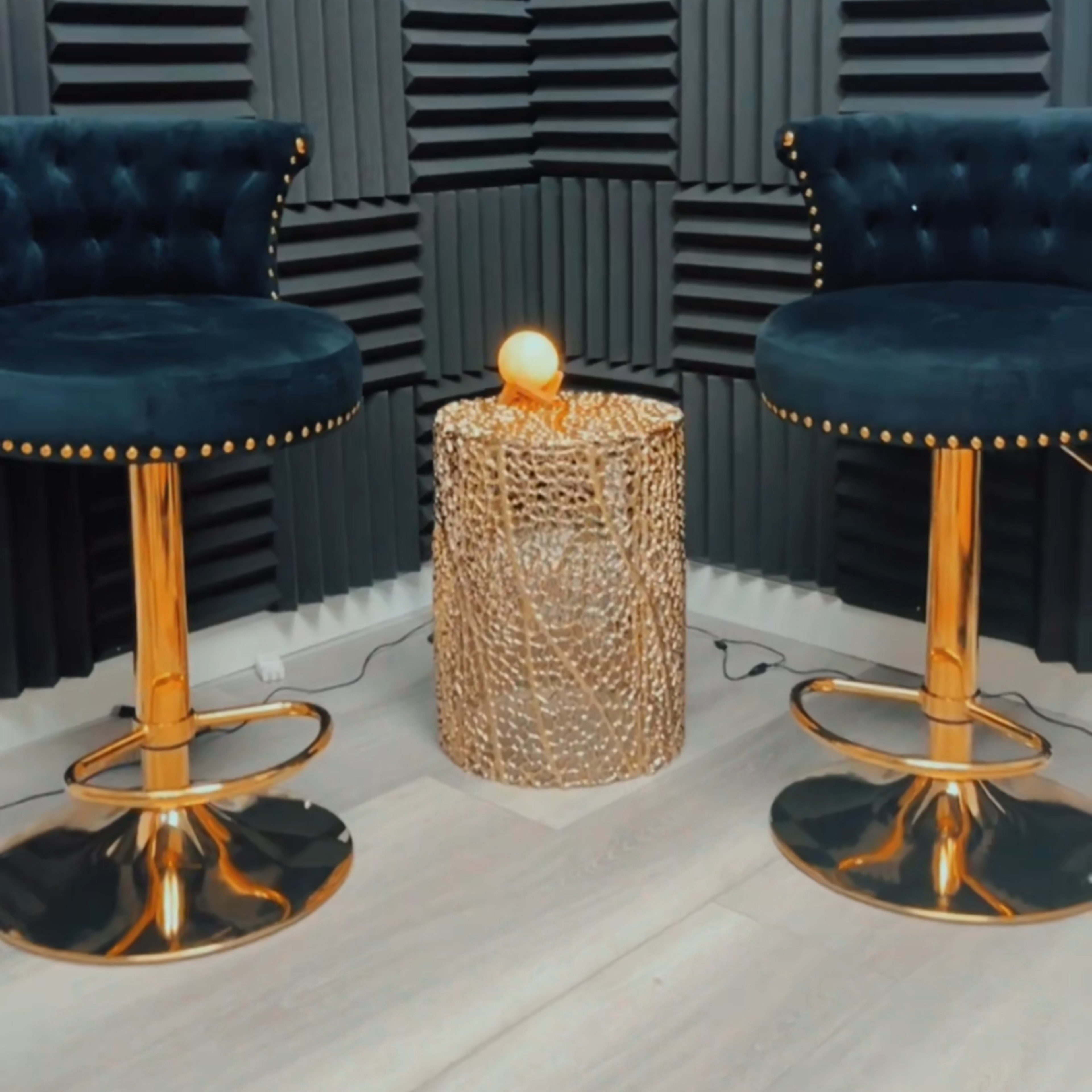 Two blue velvet bar stools with gold accents positioned on a light floor next to a woven, round table with a softly glowing orb on top, set against a wall of textured acoustic panels.