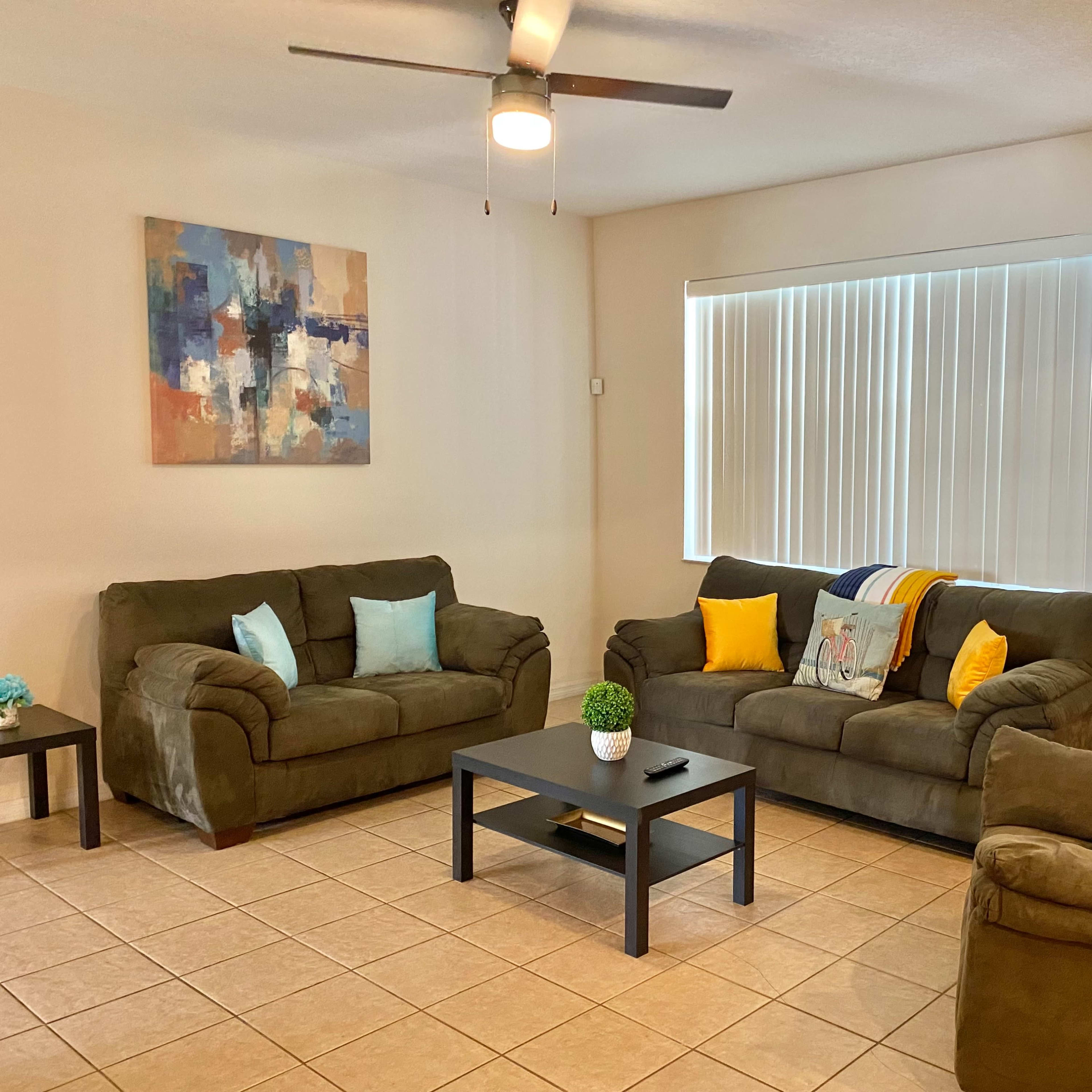 A living room features three green couches, a coffee table, a ceiling fan, and a large window with blinds.