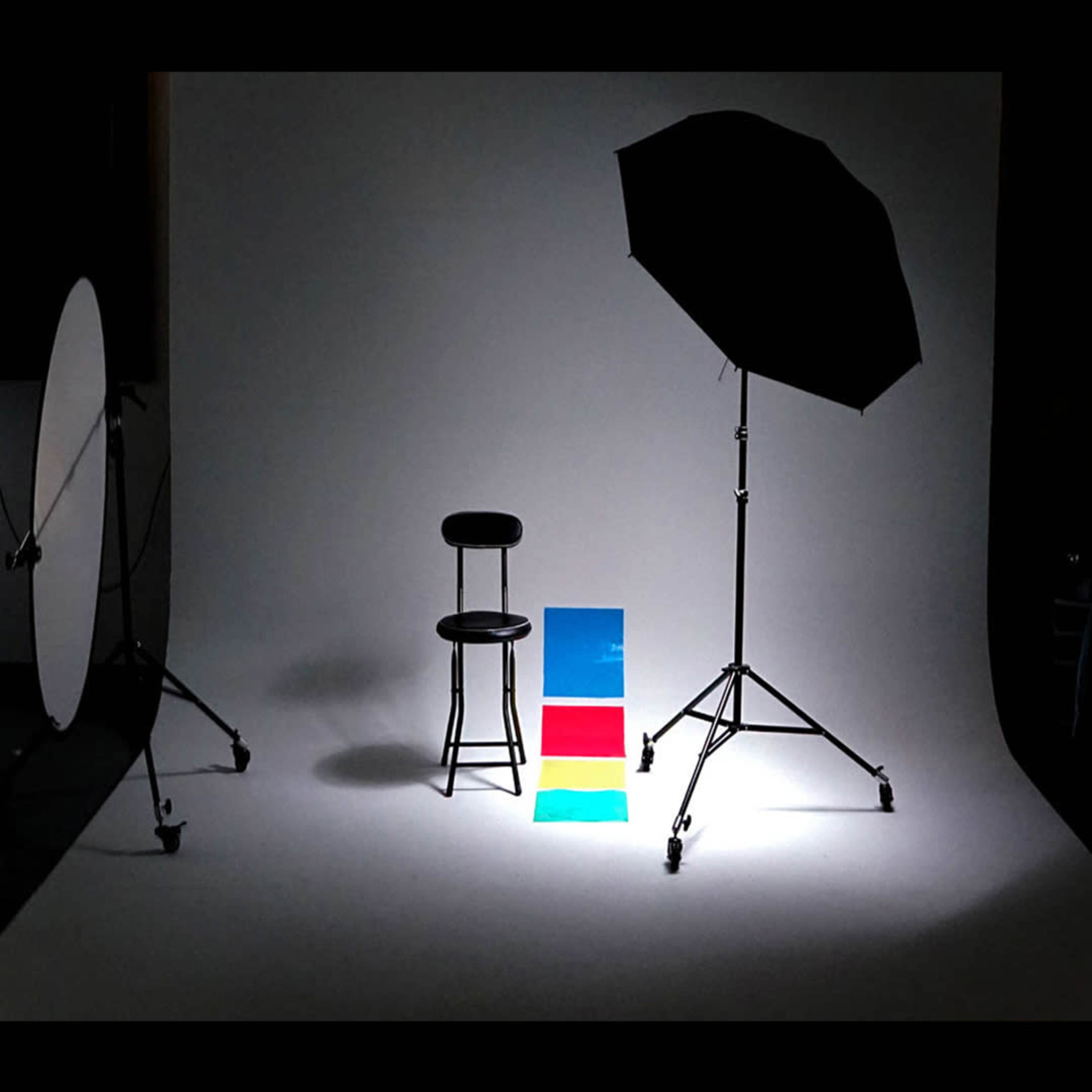 A photography setup with a black chair positioned in front of a white backdrop, along with colored panels and a large umbrella light.
