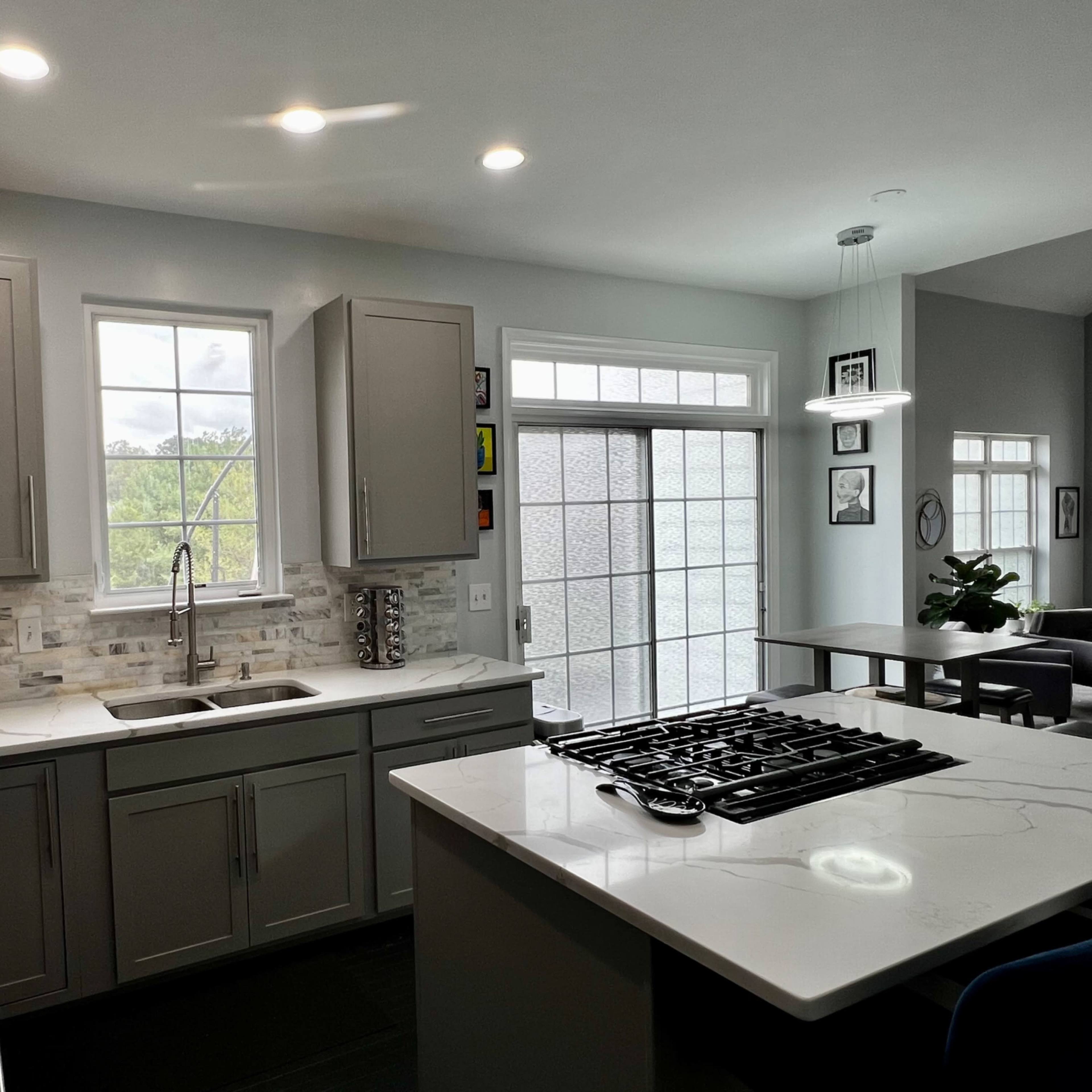 A modern kitchen features gray cabinets, a white countertop with a stovetop, and large windows that provide natural light.