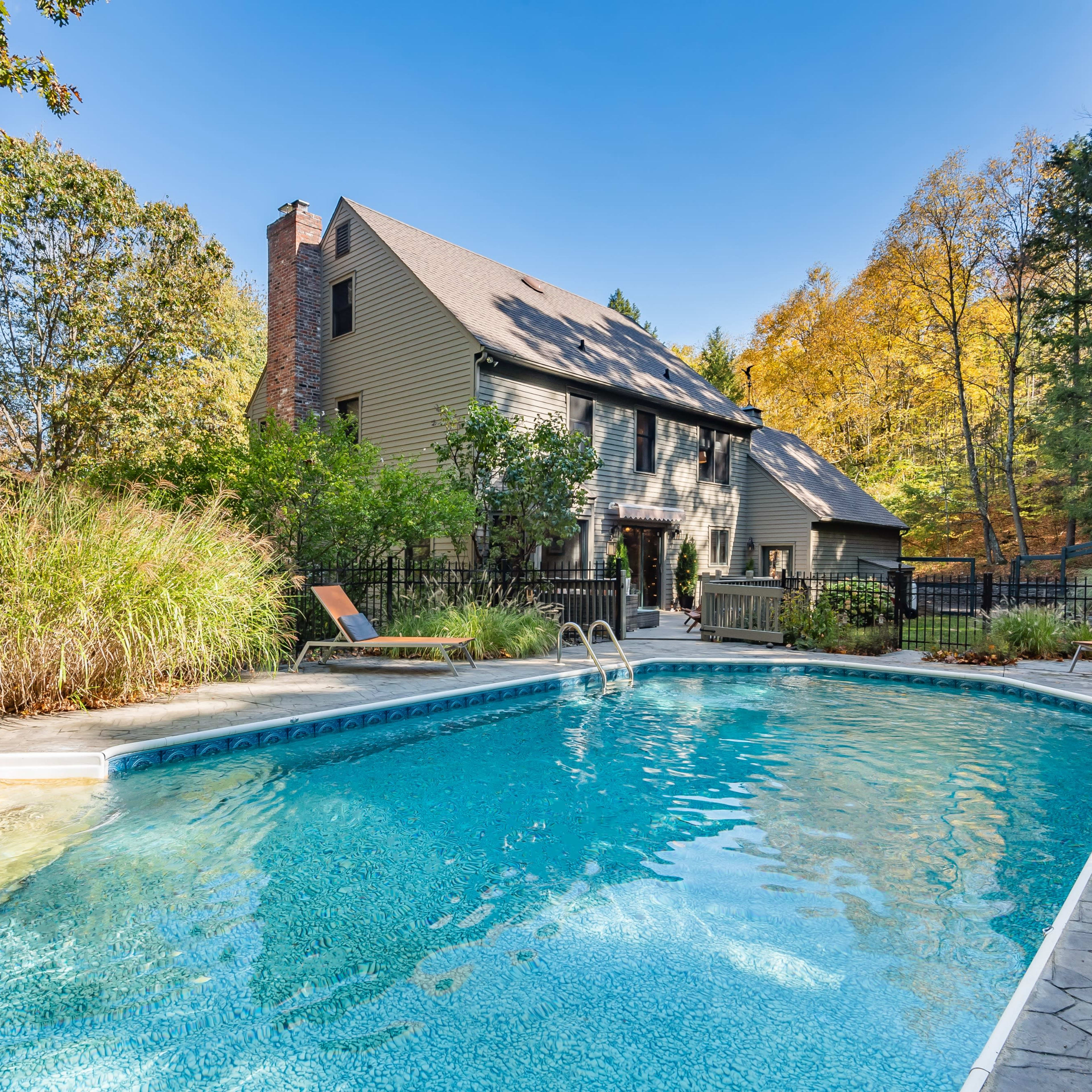 A swimming pool with a stone deck is situated next to a large house surrounded by trees in autumn foliage.