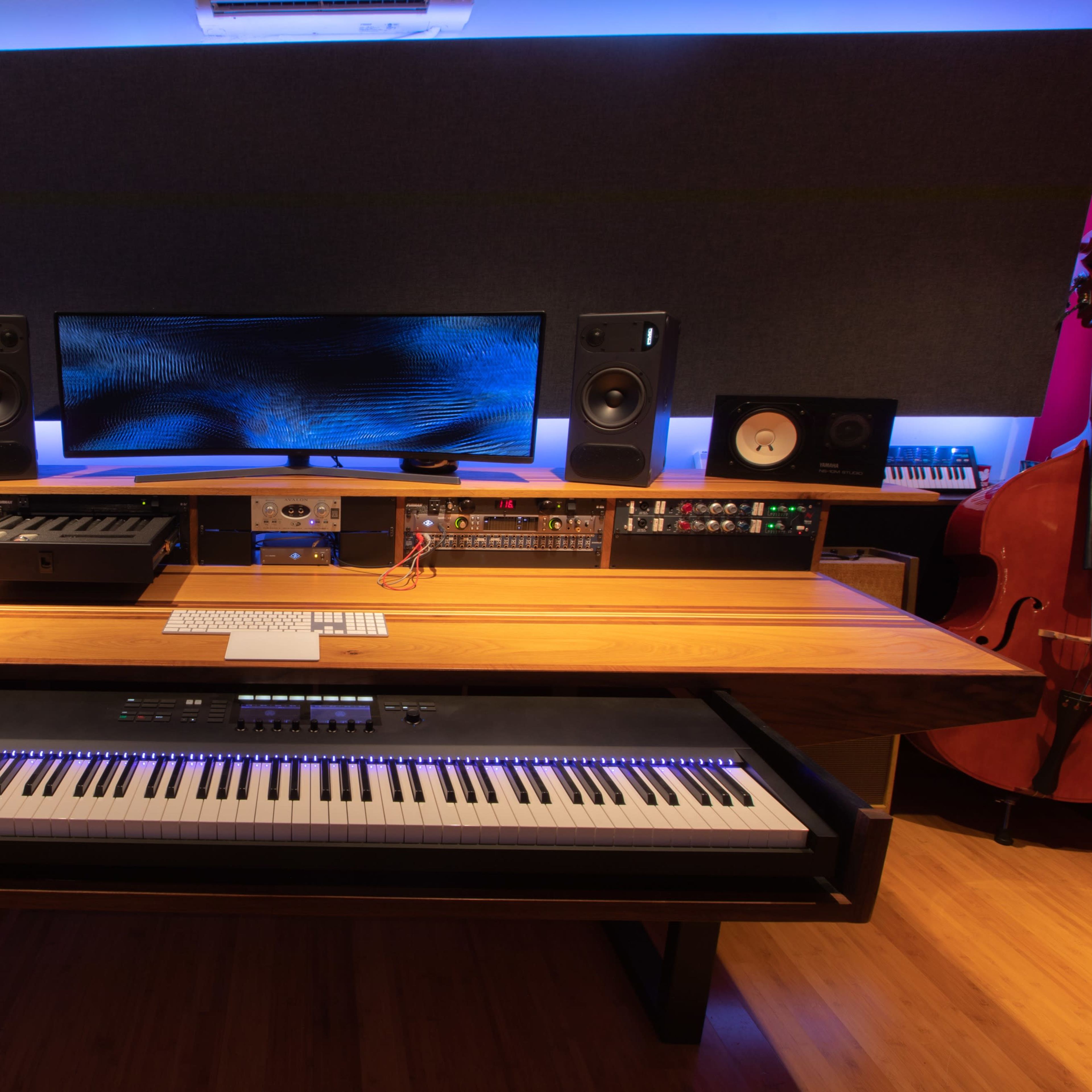 A modern music studio featuring a grand piano, audio equipment, a large monitor, and various keyboards arranged around a wooden tabletop.