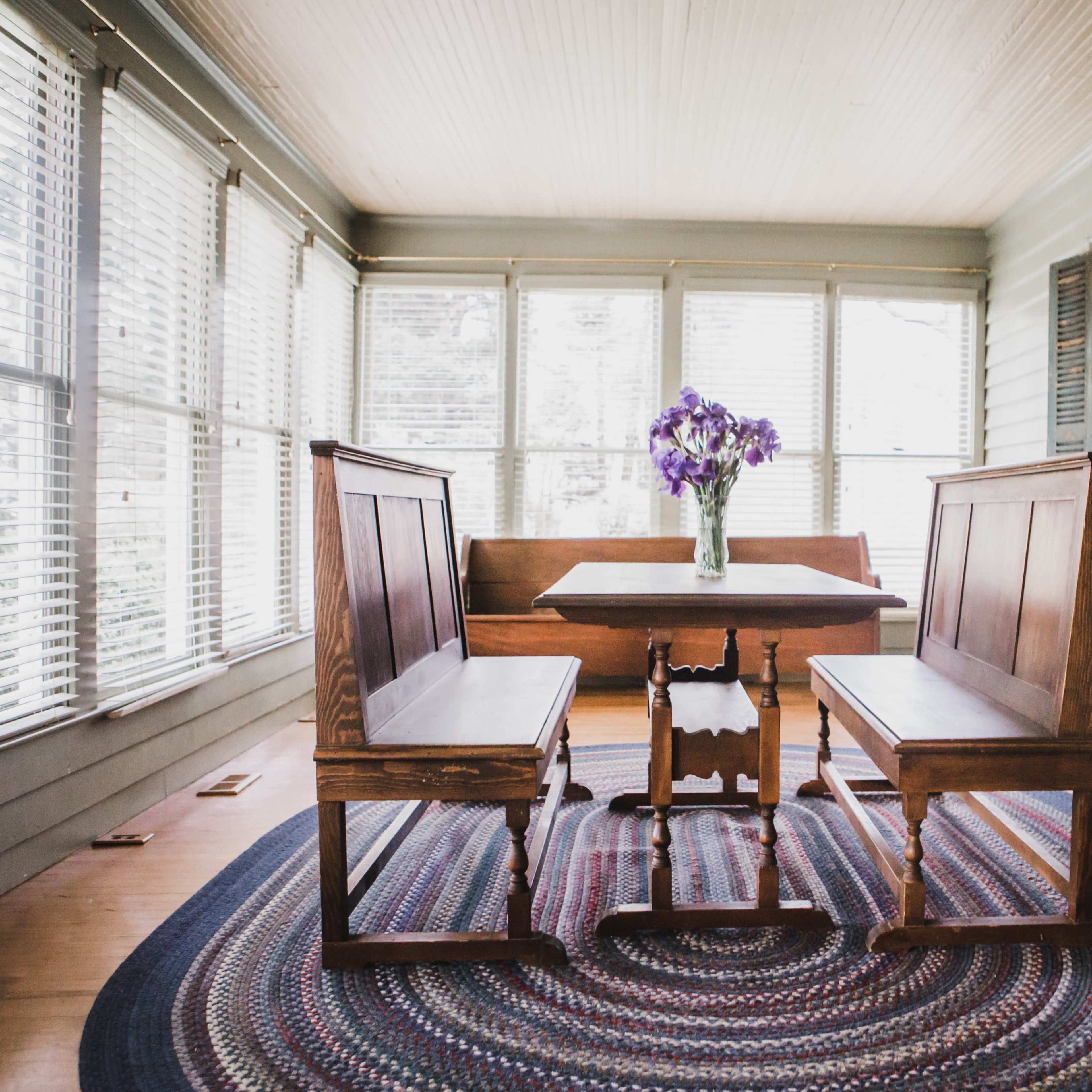 A sunlit dining area features a wooden table surrounded by two benches and a vase of purple flowers at its center.