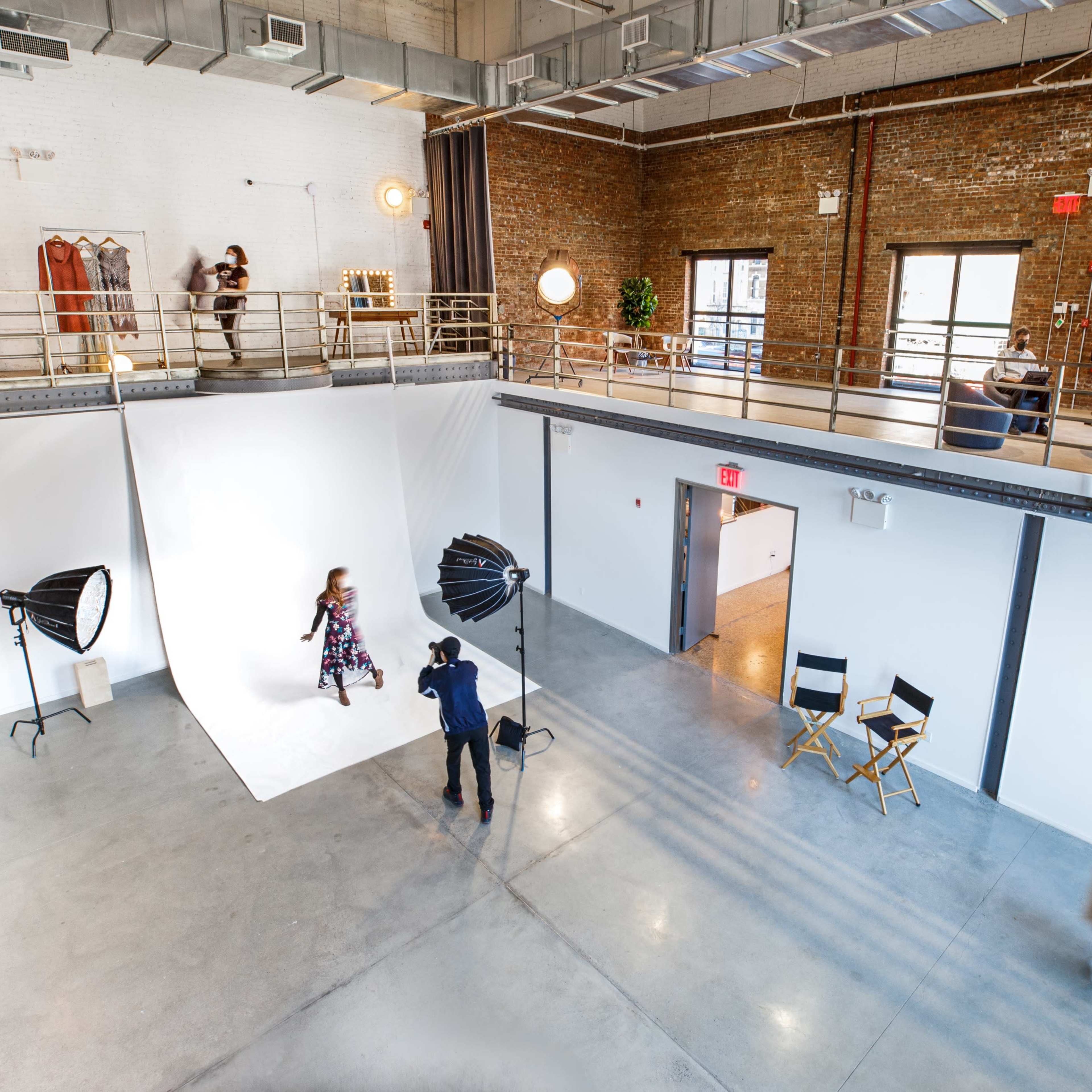 A spacious studio features a model posing on a white backdrop while a photographer captures the shot, with additional areas for changing clothes and seating visible in the background.
