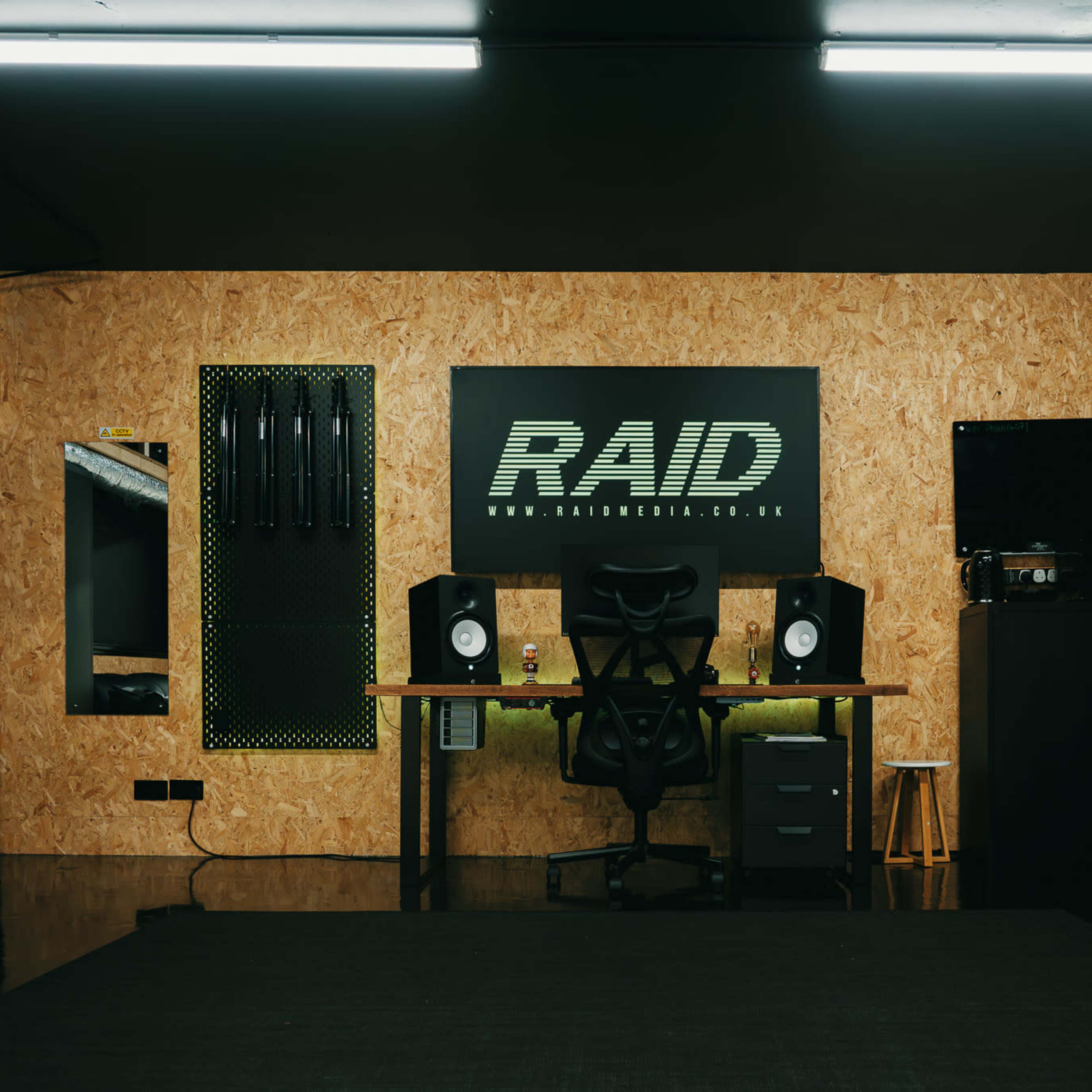 A modern workspace features a desk with audio monitors, a black chair, and a large RAID logo on an oriented strand board wall.