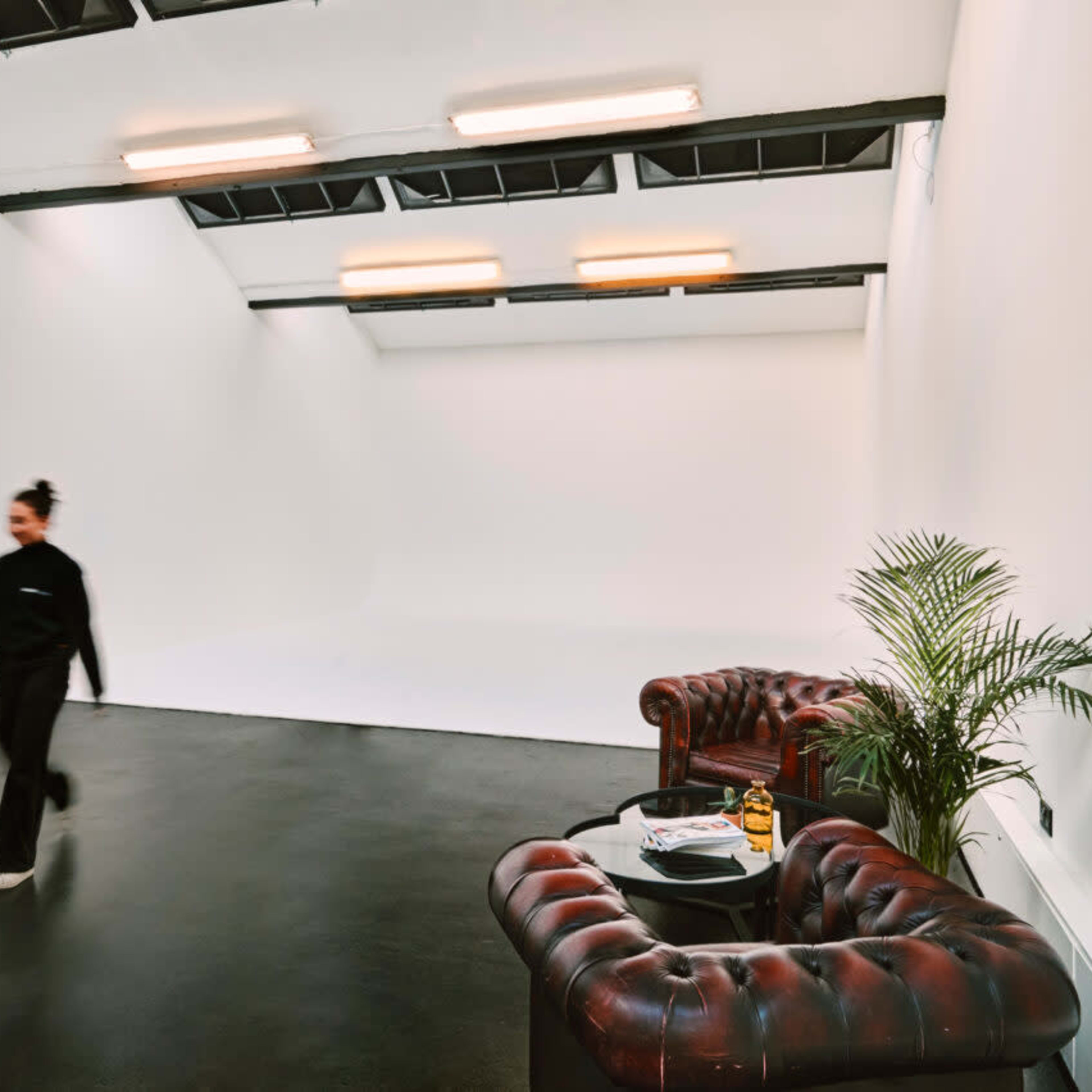 A person walks past two brown leather couches in a spacious, minimally decorated studio with a white backdrop and black flooring.