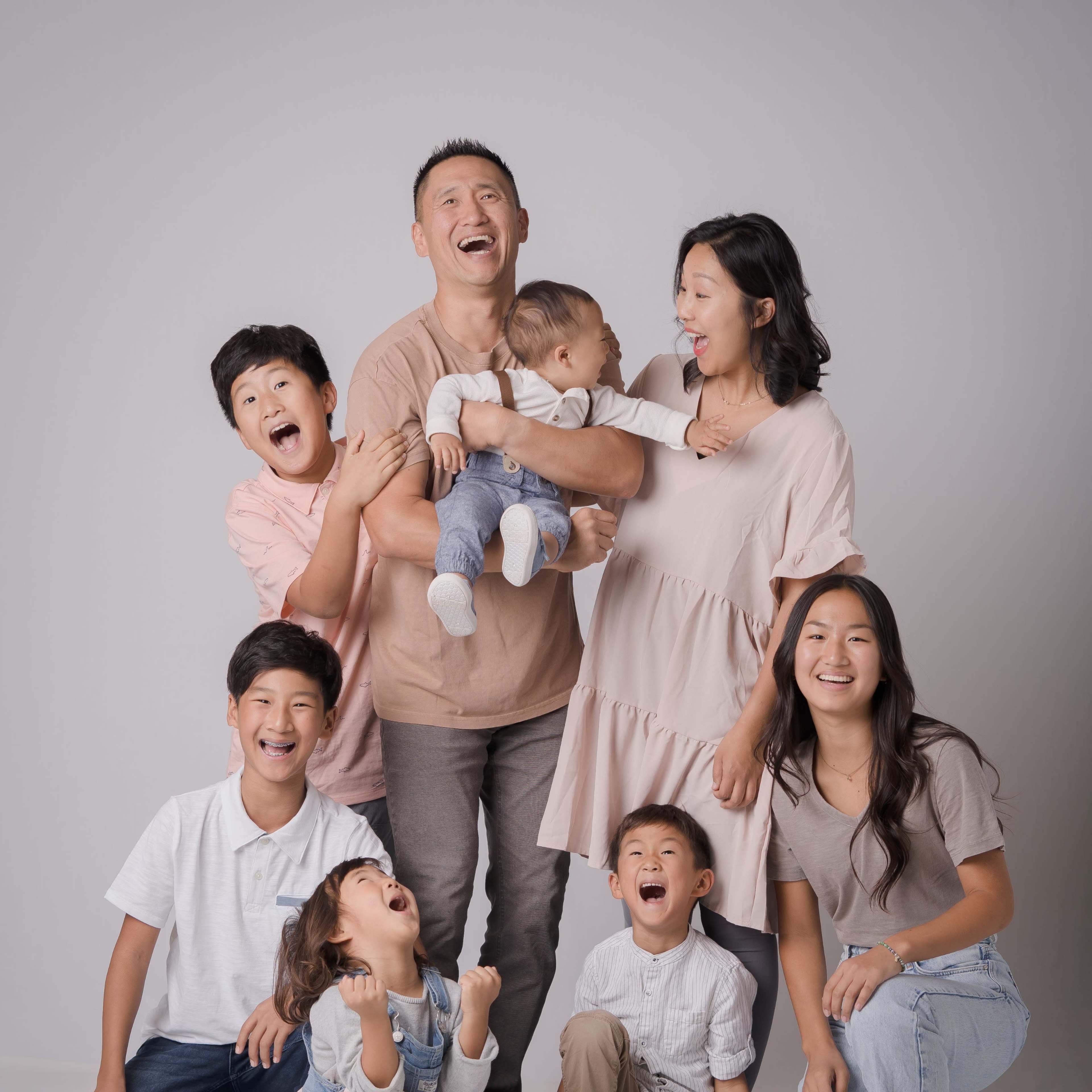 A group of eight people, including six children and two adults, are smiling and laughing together in a studio setting.