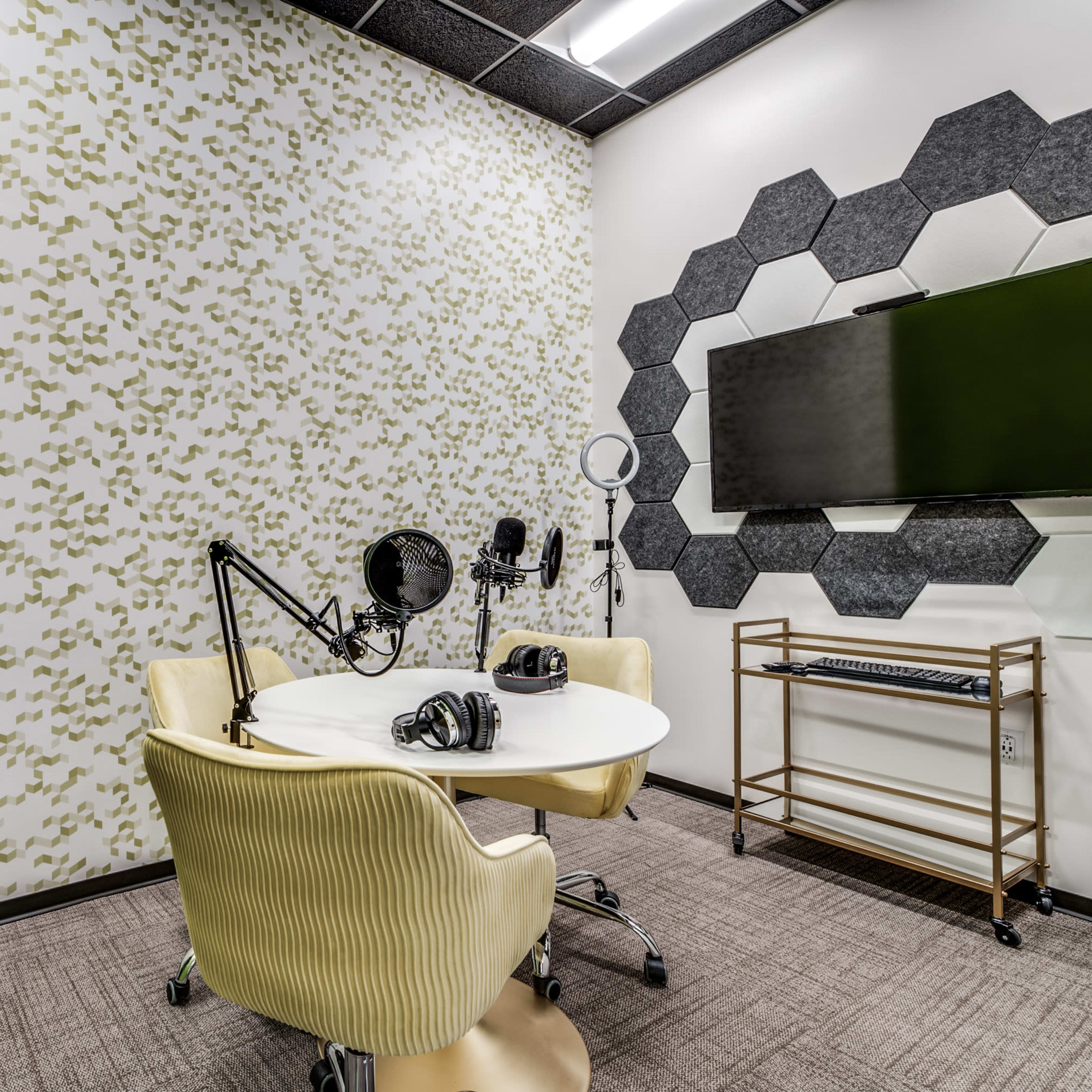 A small recording studio with a round table, two microphones, headphones, a wall-mounted screen, and a potted plant.