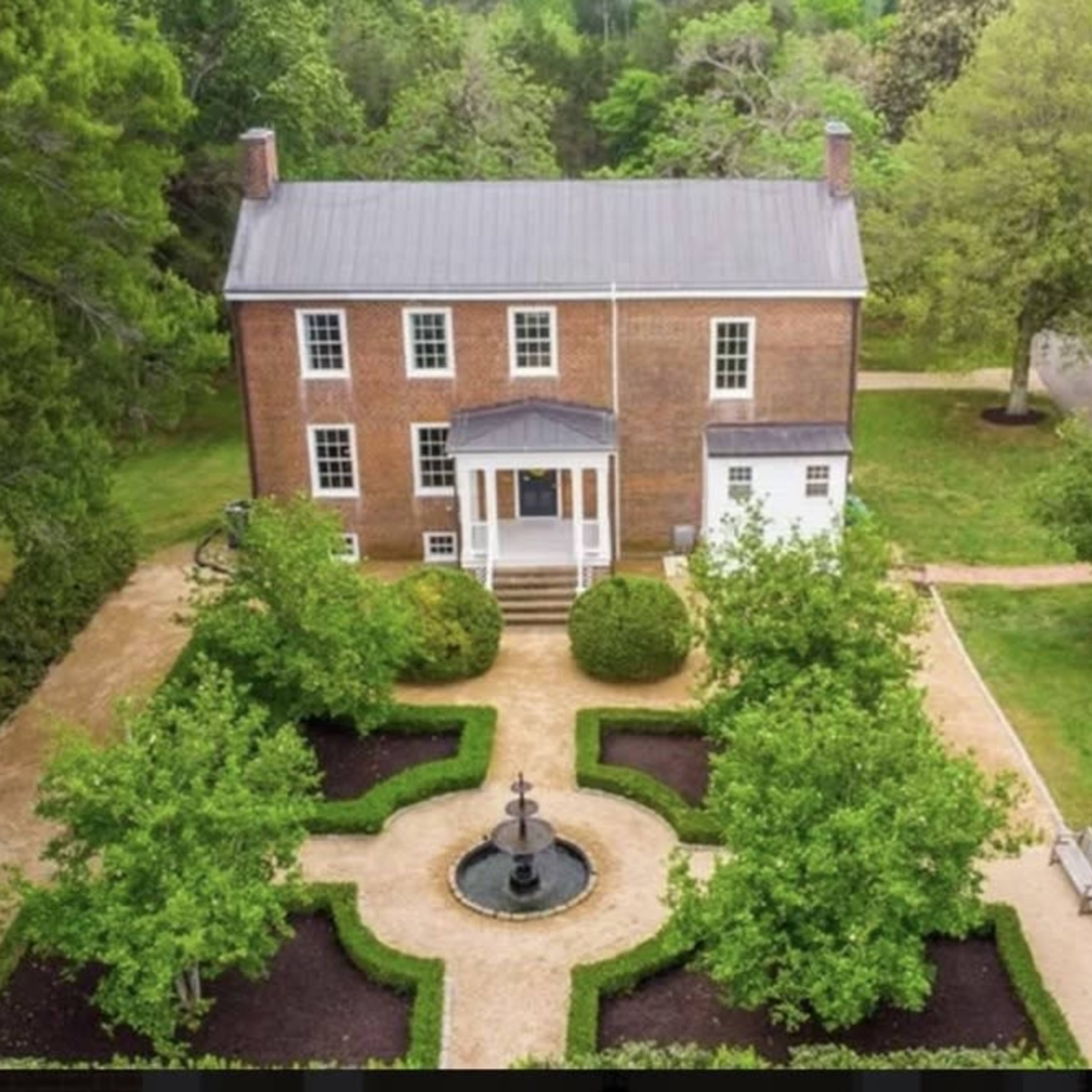 A two-story brick house surrounded by a landscaped garden featuring a central fountain and symmetrical pathways.
