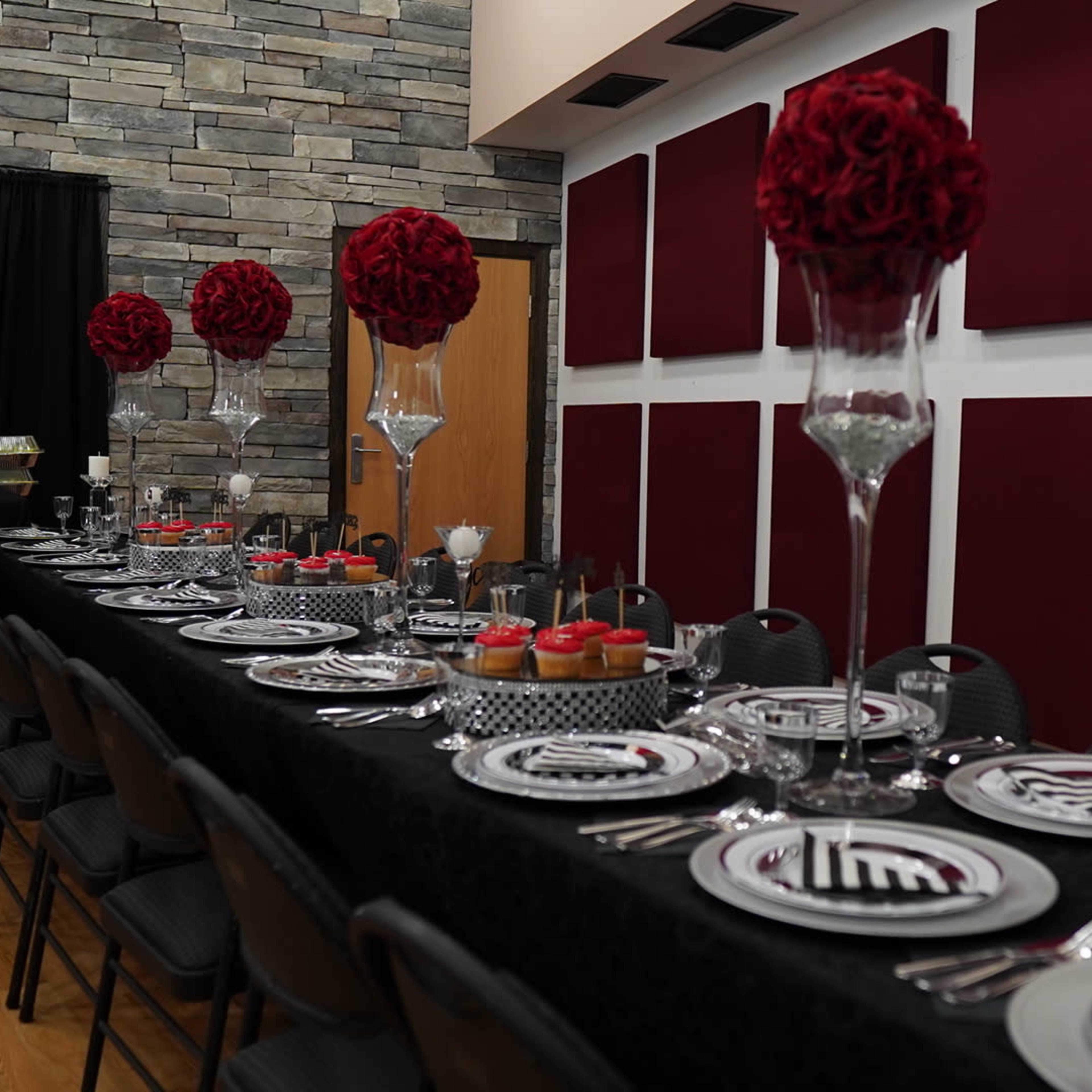 A long dining table is set with elegant tableware, floral centerpieces, and various food displays against a backdrop of stone walls and red panels.
