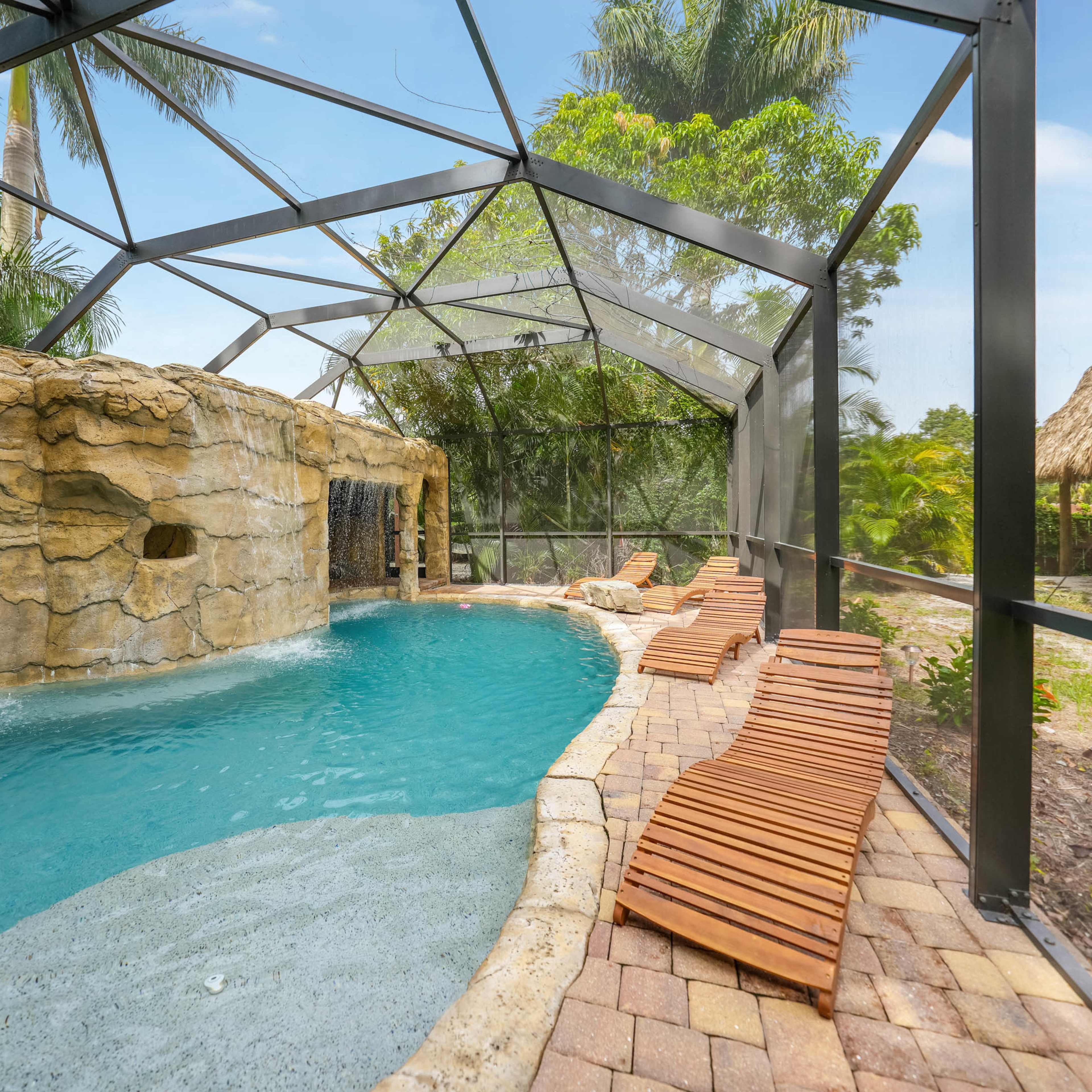 A pool area enclosed by a clear mesh screen, featuring a rock waterfall and wooden lounge chairs beside the water.