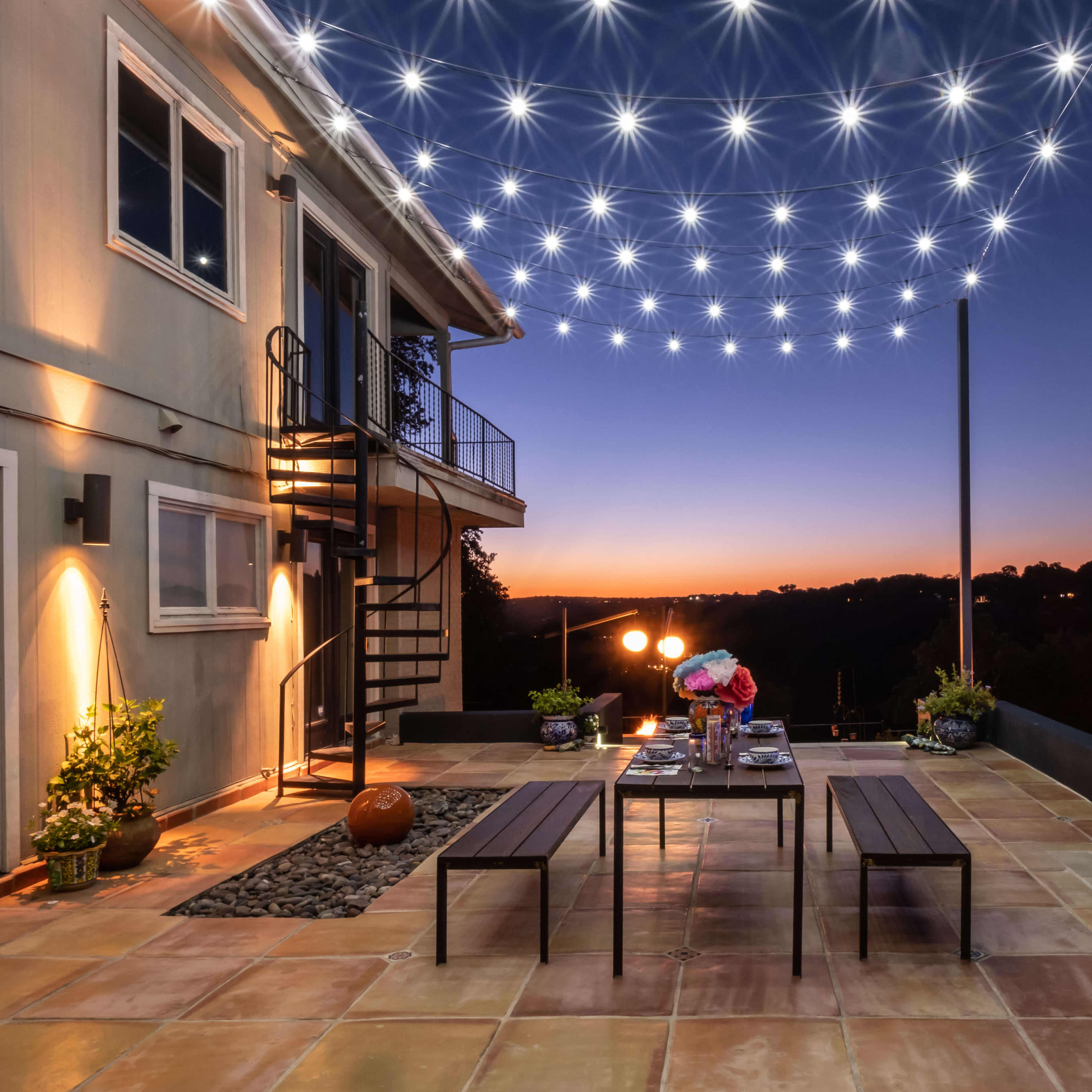 A patio with a dining setup is illuminated by string lights at dusk, featuring a pool and a fire feature in the background.