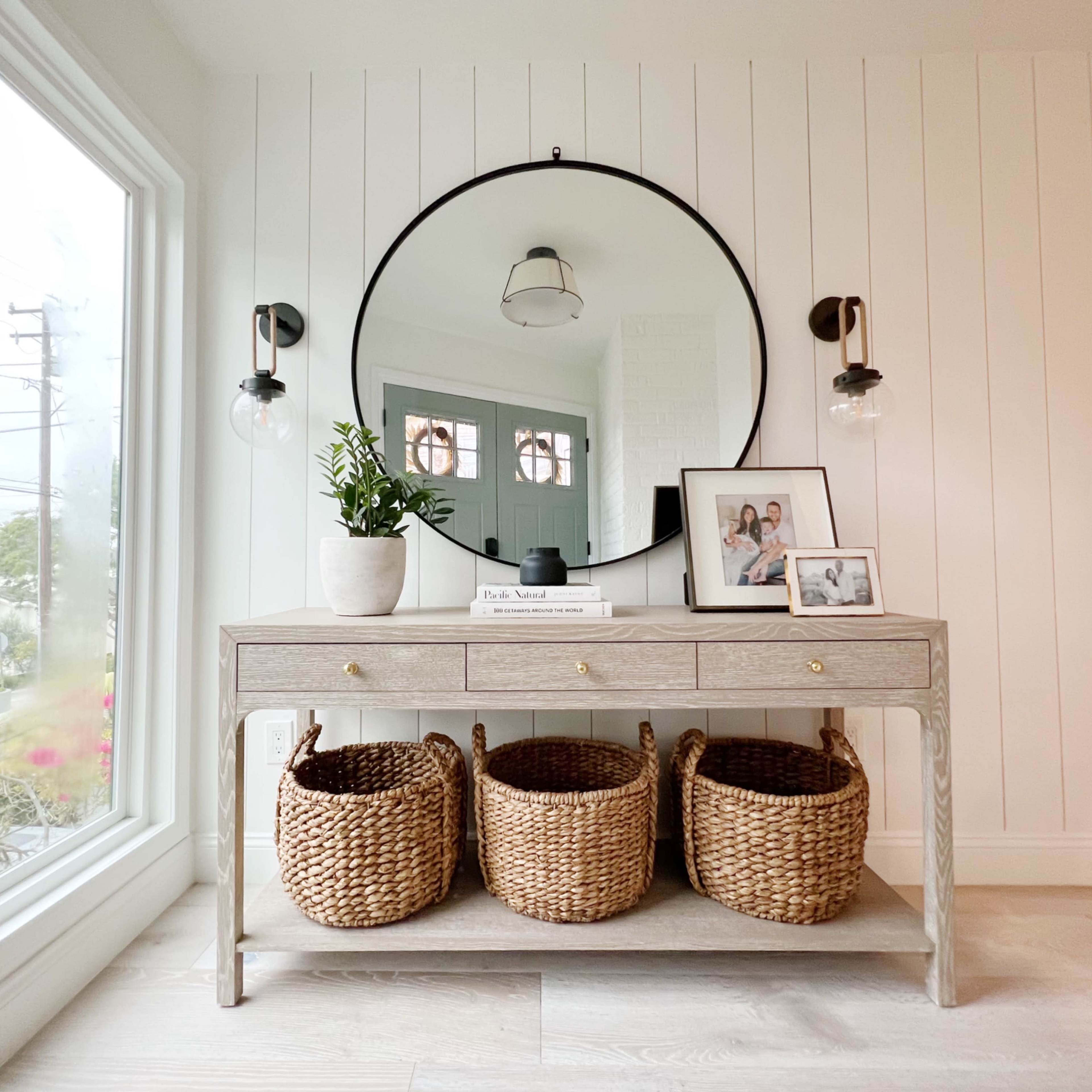 A wooden console table with three woven baskets sits beneath a large round mirror, illuminated by wall sconces, in a bright room with shiplap walls and a window view.