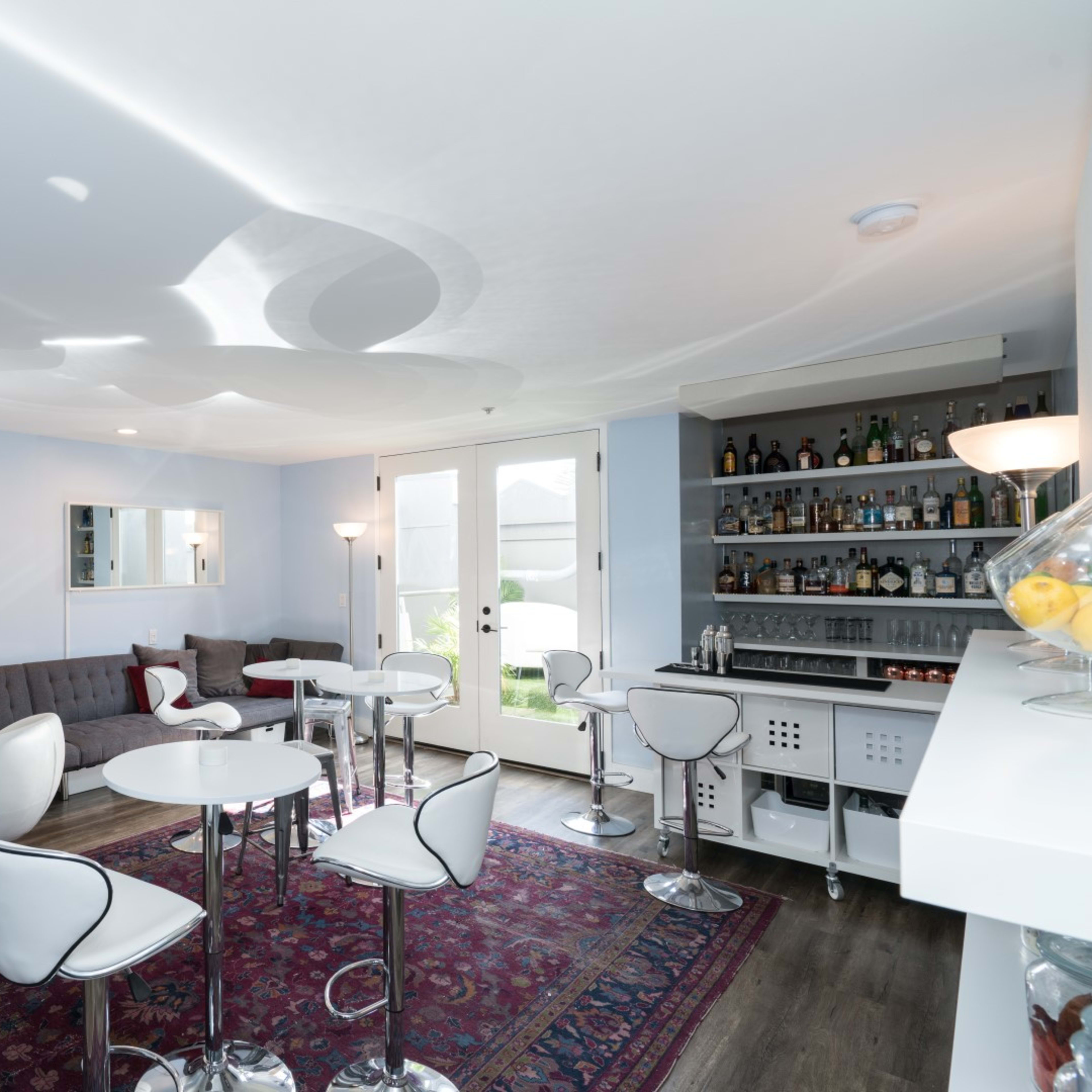 A modern lounge area features a gray sectional sofa, white bar stools, circular tables, and a well-stocked bar on one side.